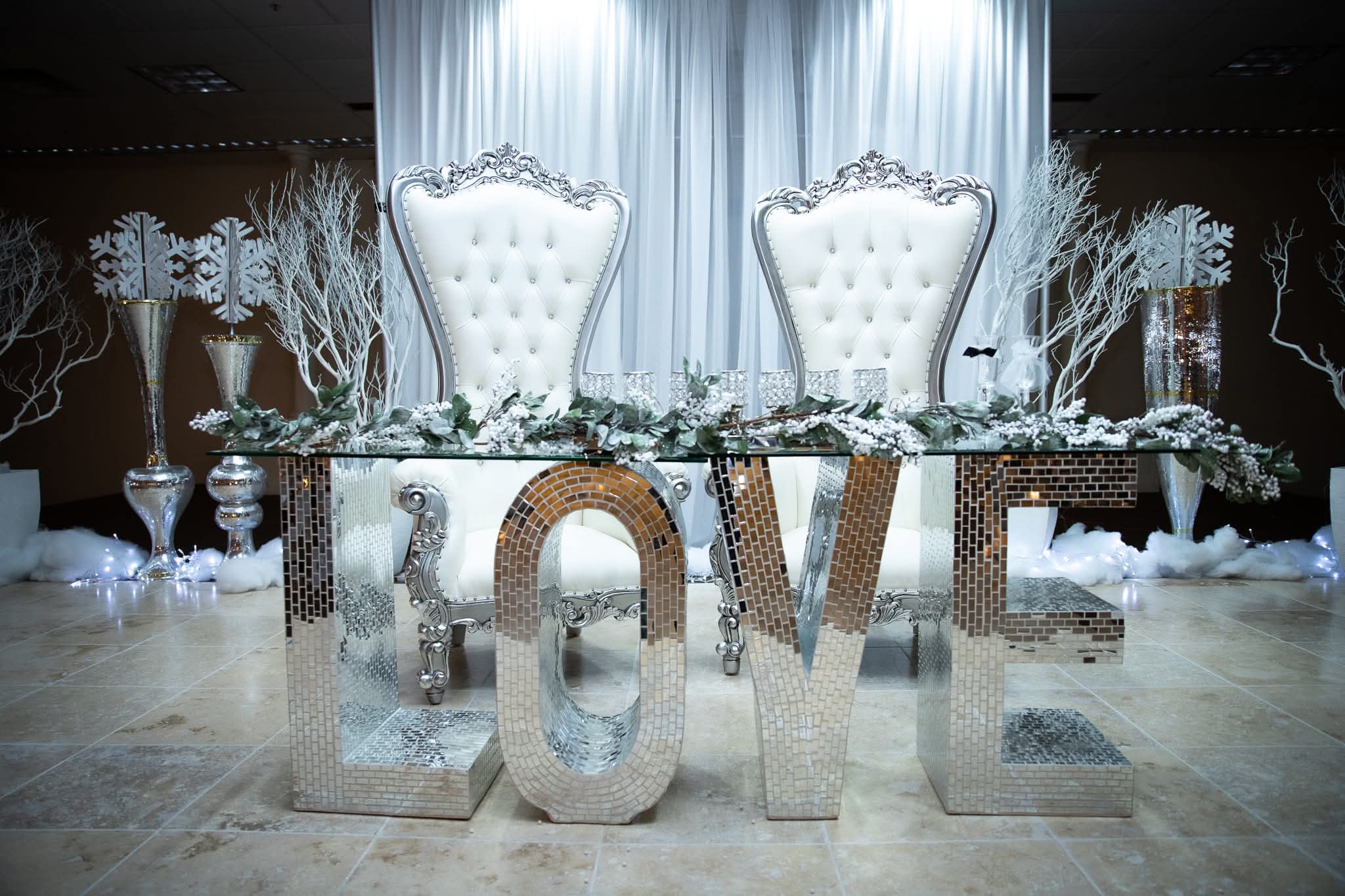Two ornate white chairs behind a table with a floral arrangement, with large reflective