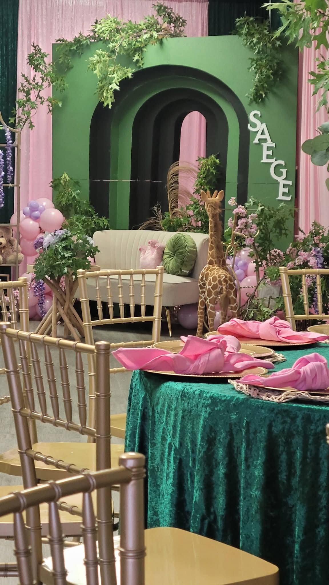 Decorative setting with a green backdrop, pink and purple accents, a table with pink napkins, and chairs, including a gold one.
