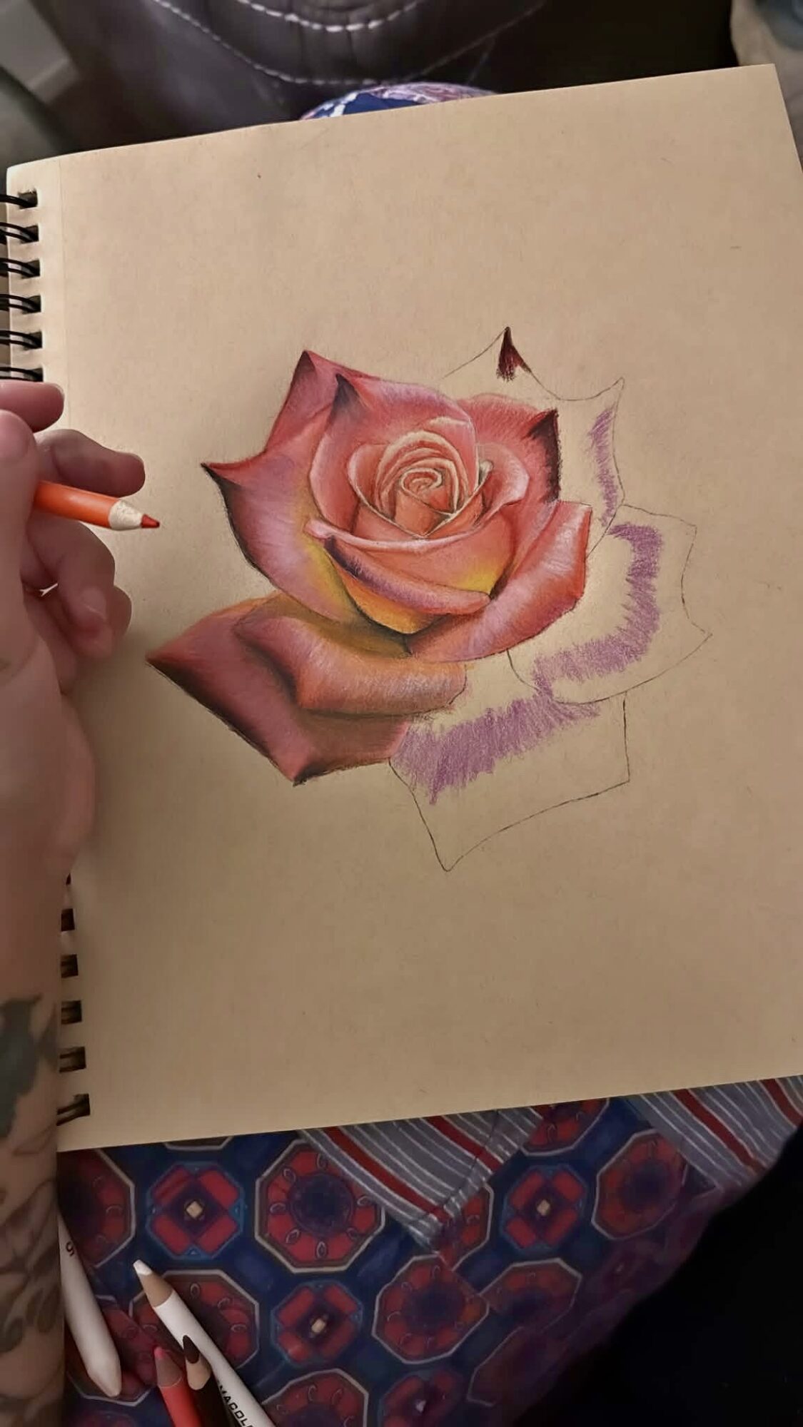 Hand holding a pencil drawing a pink rose on paper, with a partially completed sketch and colored petals.