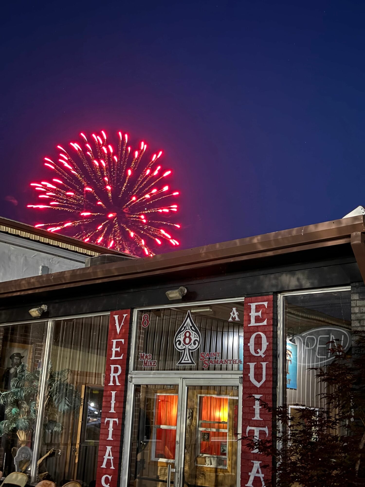 Fireworks explode in the night sky above a building with a sign, illuminated windows visible, and a tree nearby.