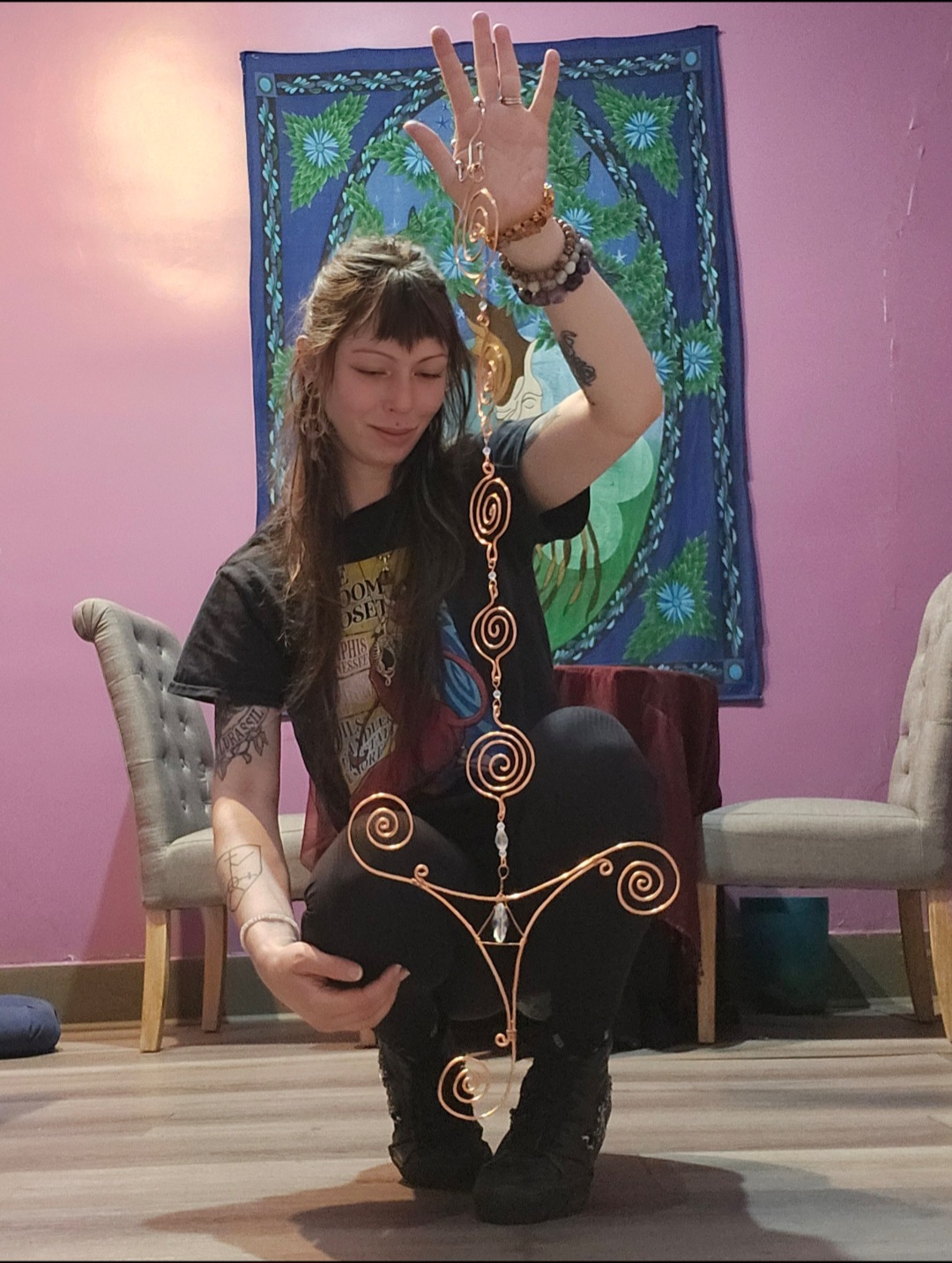 Young woman kneeling with one hand raised, colorful jewelry, in a room with pink walls and decorative tapestry.