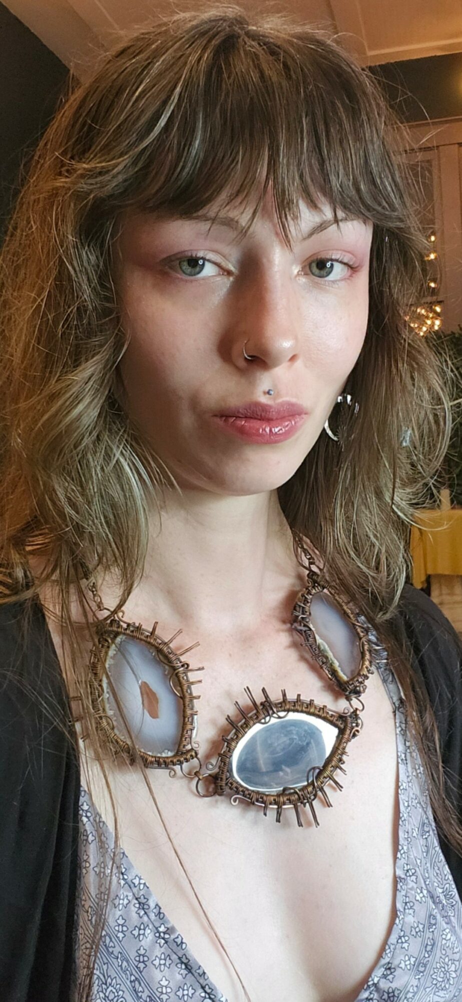Young woman with wavy hair wearing a large statement necklace with blue stones, indoors with warm lighting.