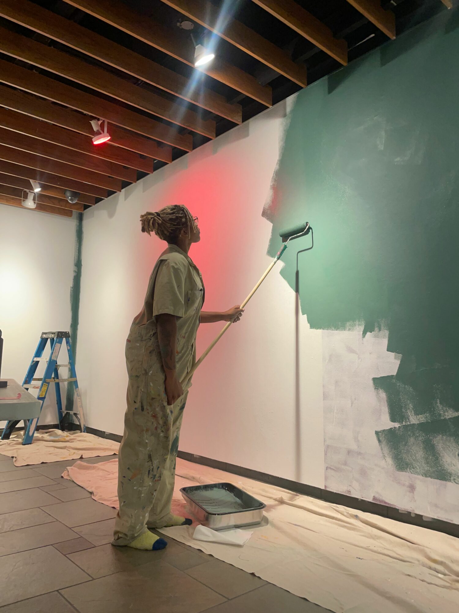 Person painting a wall with a roller, standing on the floor, with painting supplies nearby.