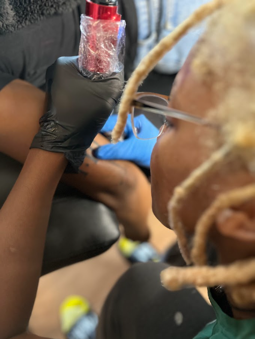 Person with glasses and dreadlocks receives a tattoo from an artist wearing gloves, holding a tattoo machine and ink container.