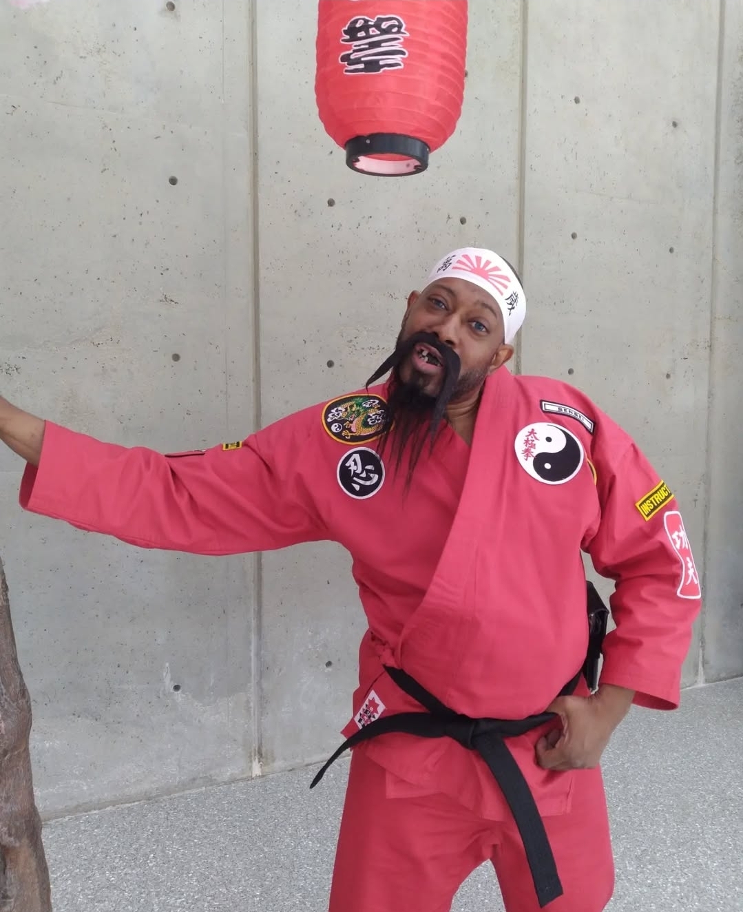 Person in a red martial arts uniform with patches, standing against a concrete wall, holding a red paper lantern above head.