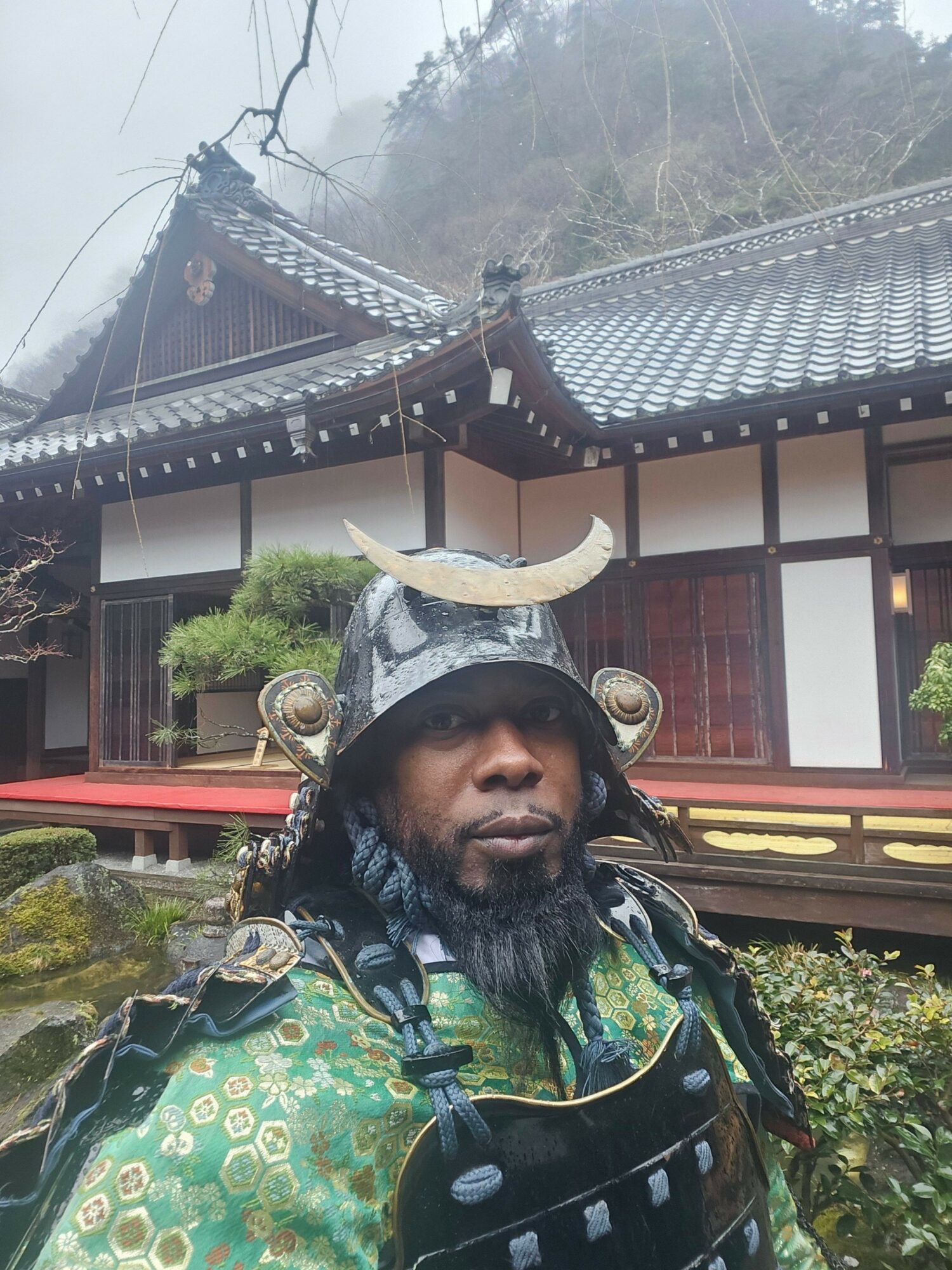 Person in traditional attire with a helmet, standing outdoors in front of a building with a tiled roof.