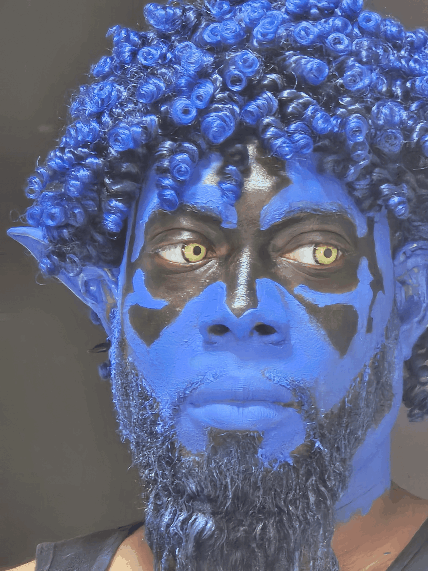 Person with blue face paint, curly blue hair, and yellow eyes, looking to the side.