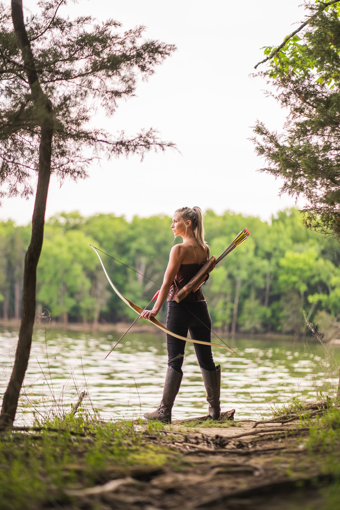 Woman standing by a lake holding a bow and arrow, surrounded by trees and greenery.
