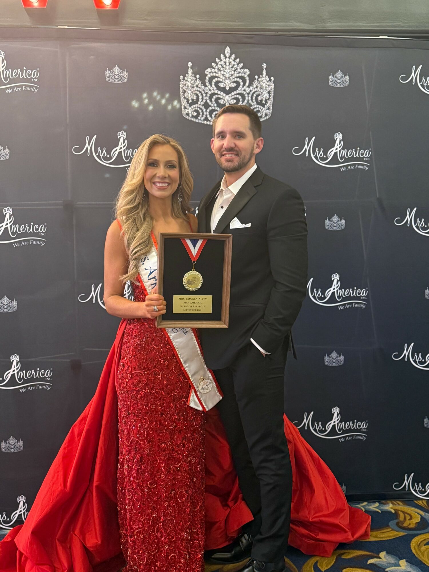 A woman in a red gown holding a medal, standing next to a man in a black suit, in front of a backdrop with logos.