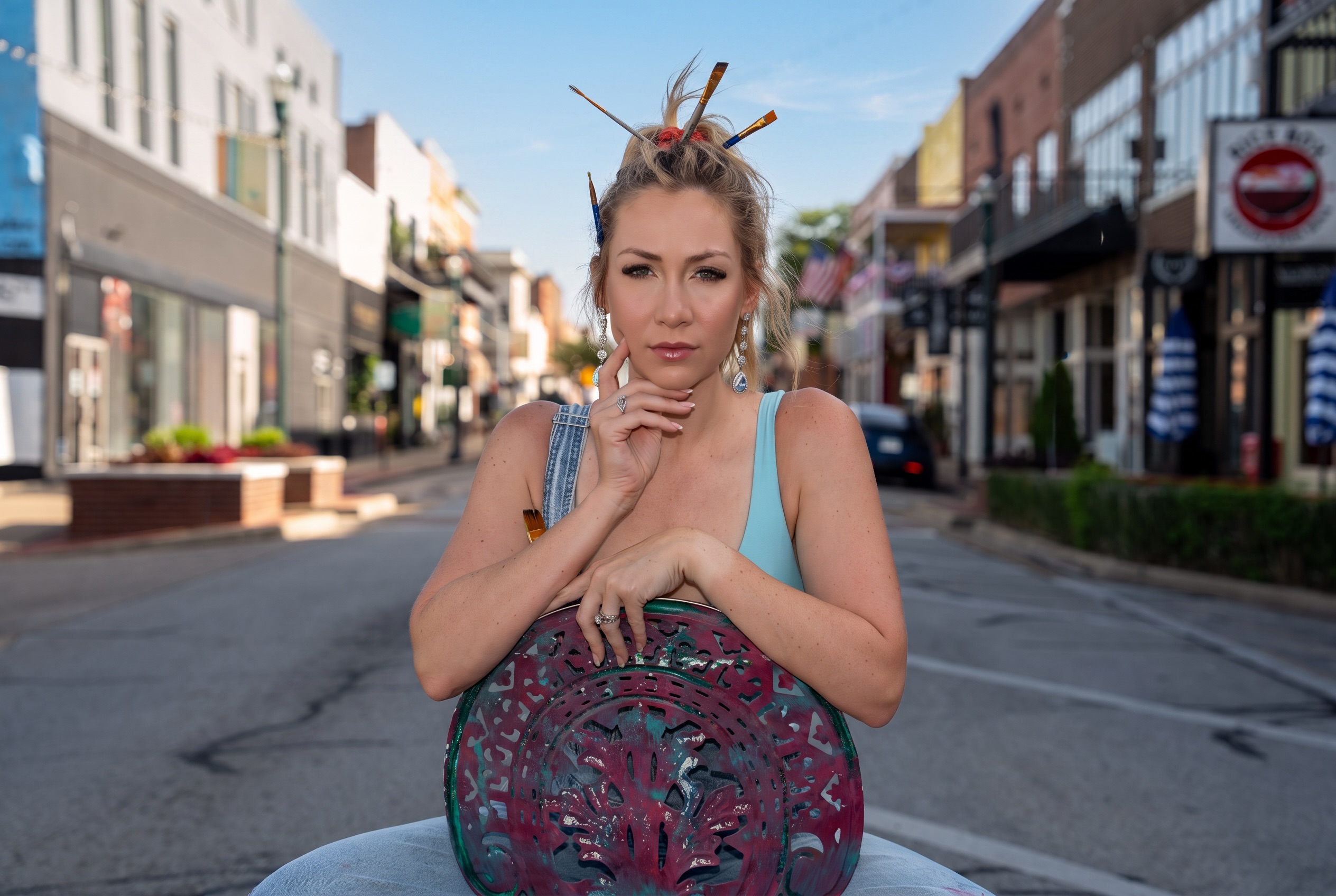 Woman with styled hair and makeup, sitting outdoors on a city street, holding a decorative object, looking at the camera.