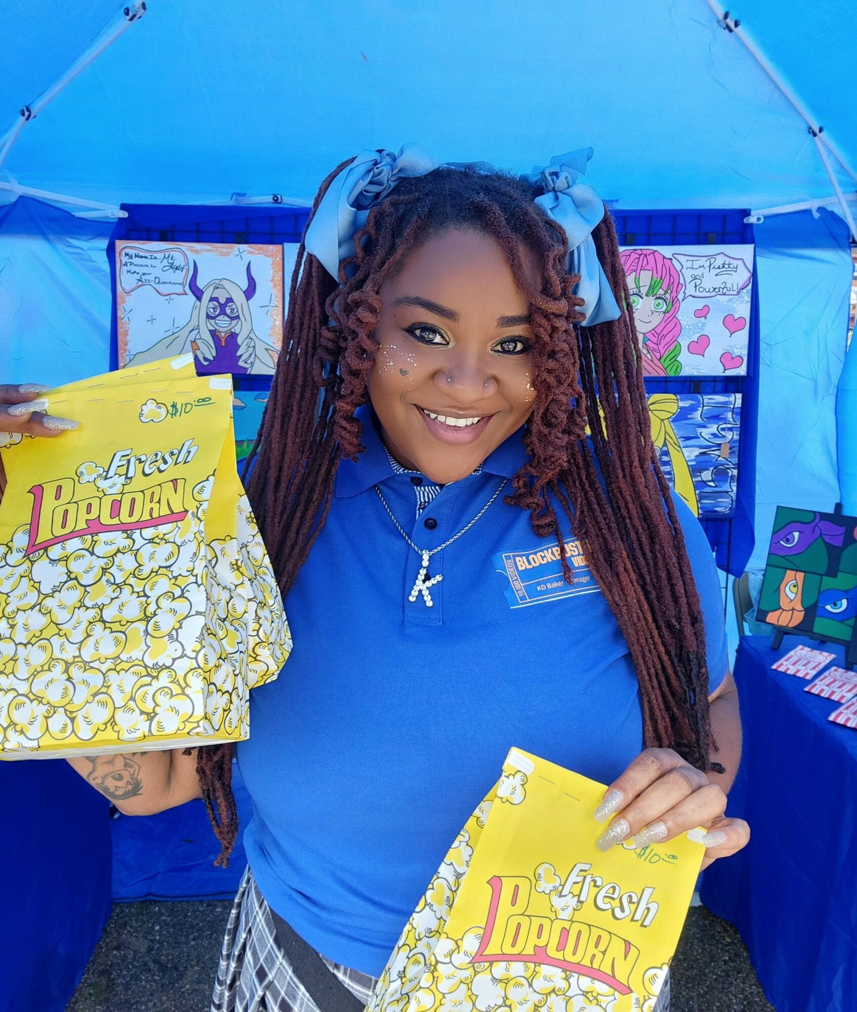 Smiling woman with braided hair in a blue shirt holding yellow popcorn bag at a fair or event.