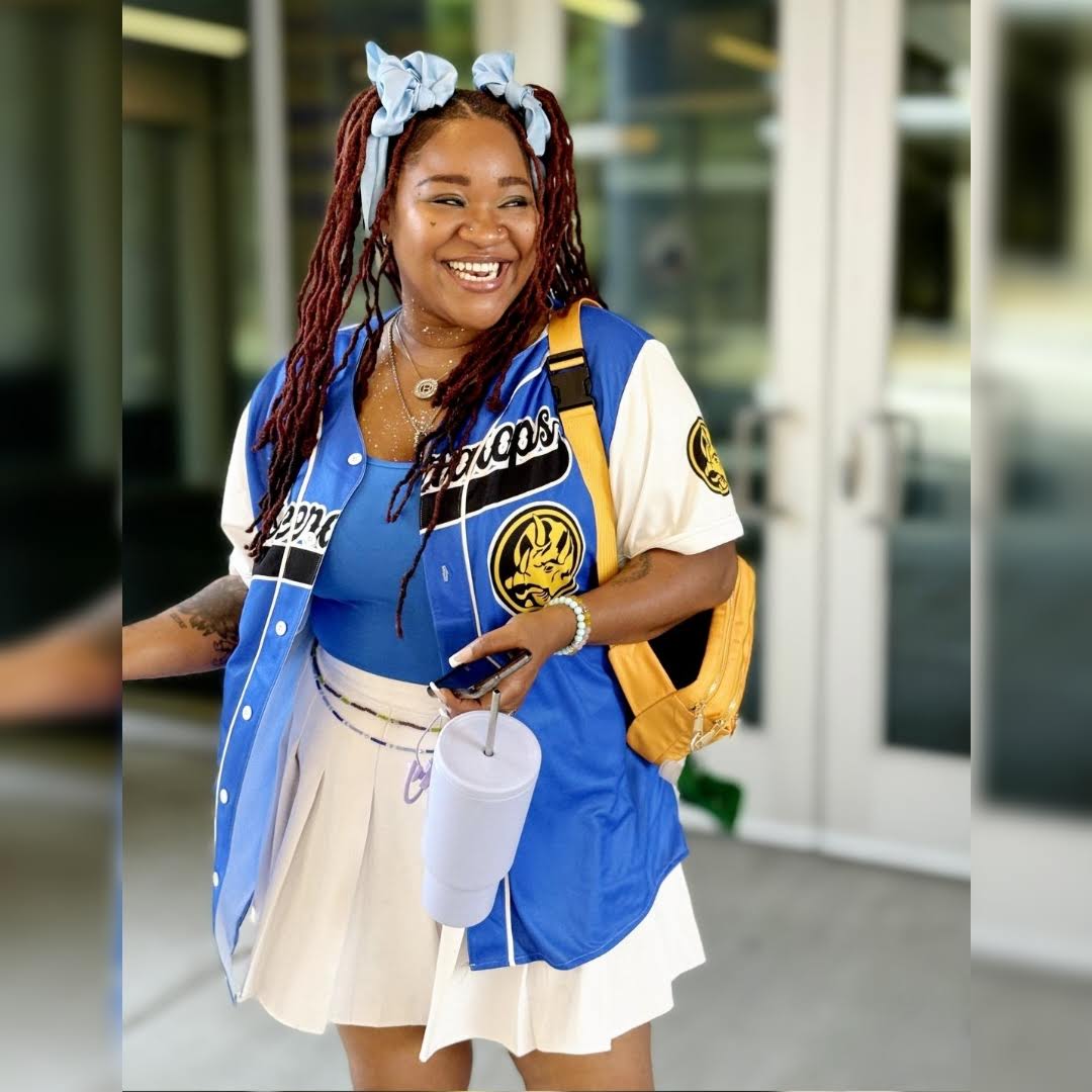 Smiling woman with a blue and white outfit, carrying a yellow backpack and holding a drink and phone, standing outside.