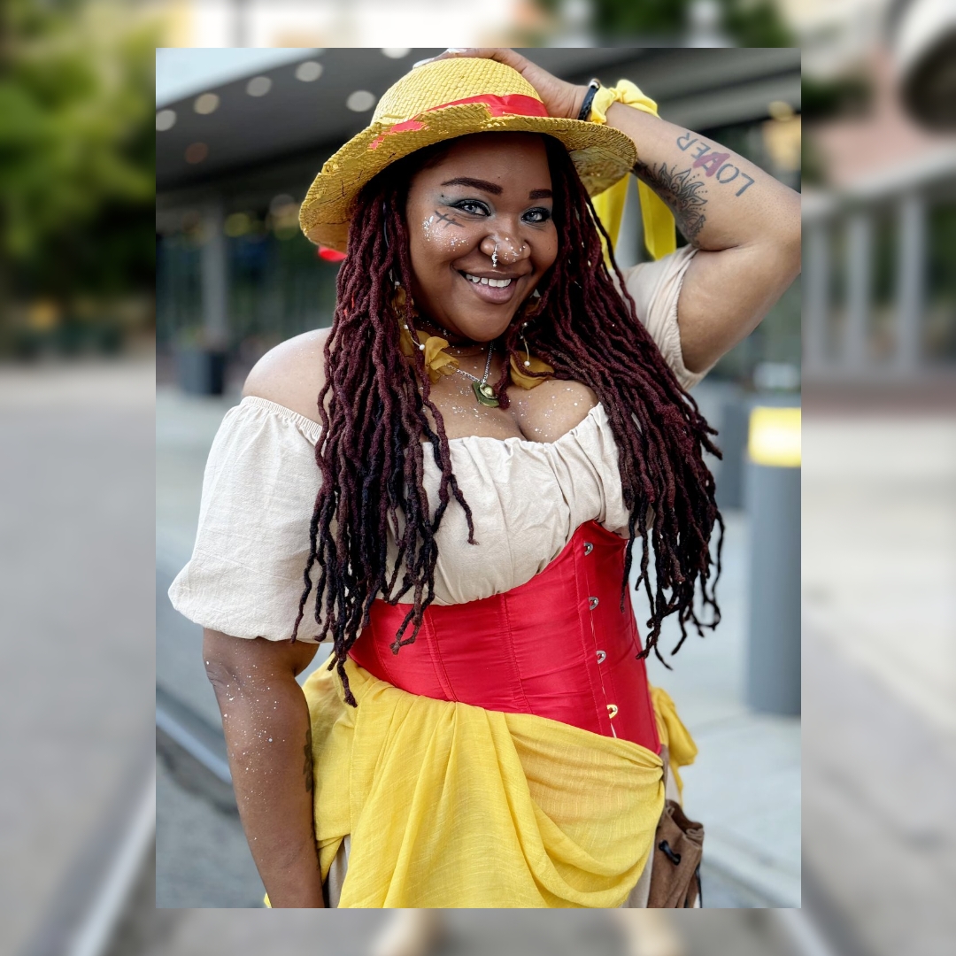 Smiling woman with dreadlocks wearing a straw hat, off-shoulder top, red corset, and yellow skirt outdoors.