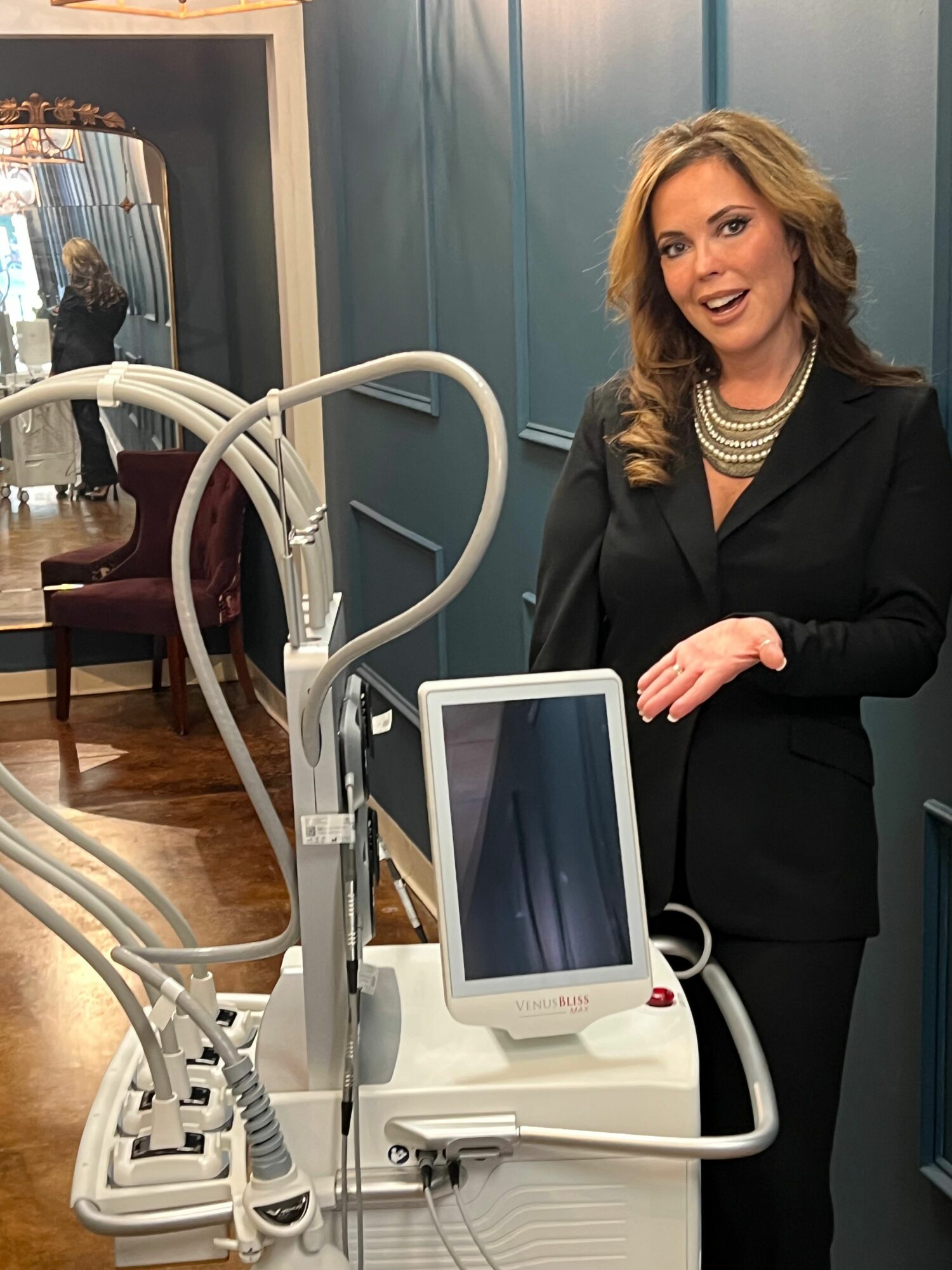 Woman in black suit gesturing next to medical equipment in a clinical setting.