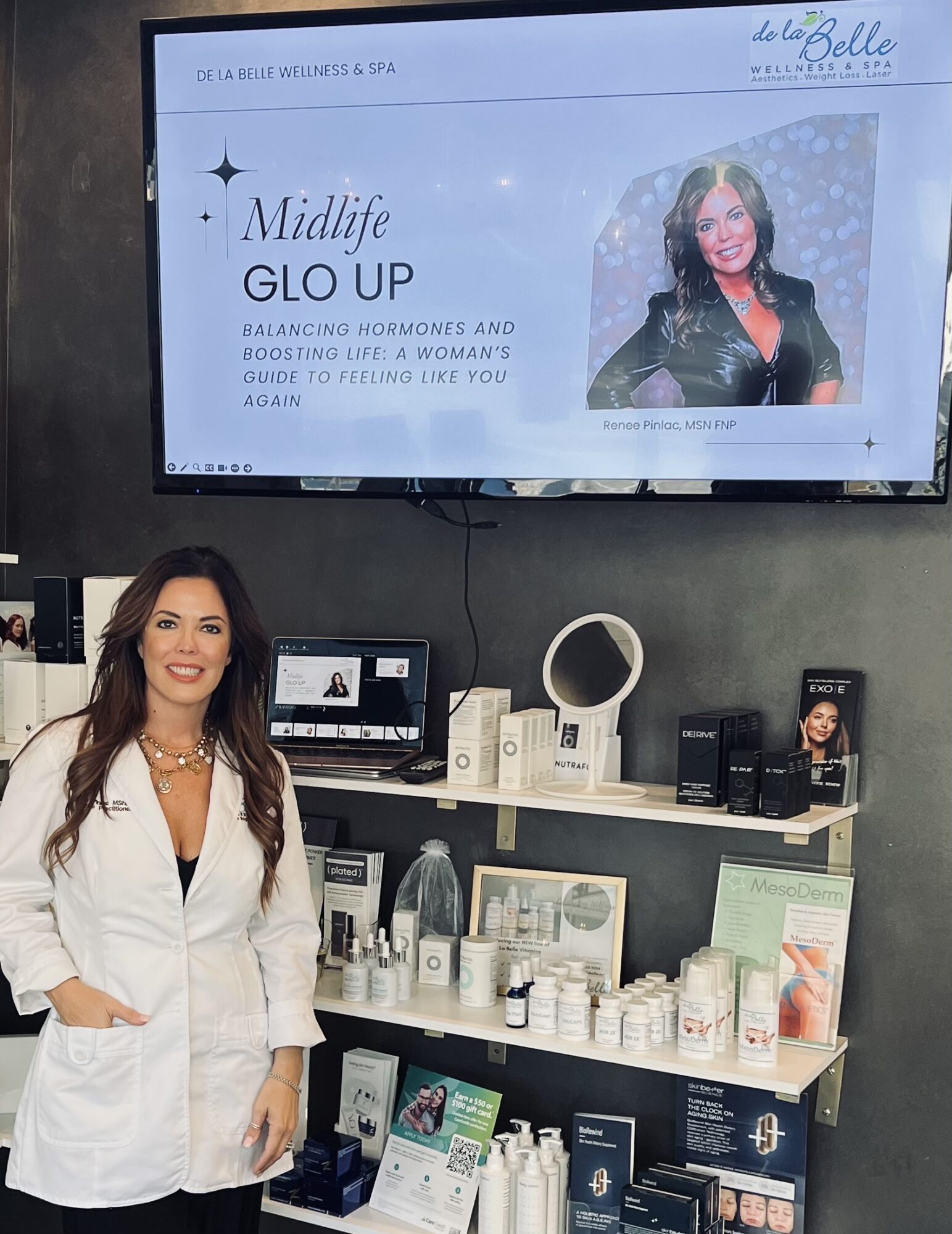 Woman in white coat standing in front of shelves with skincare products and a display screen behind her.