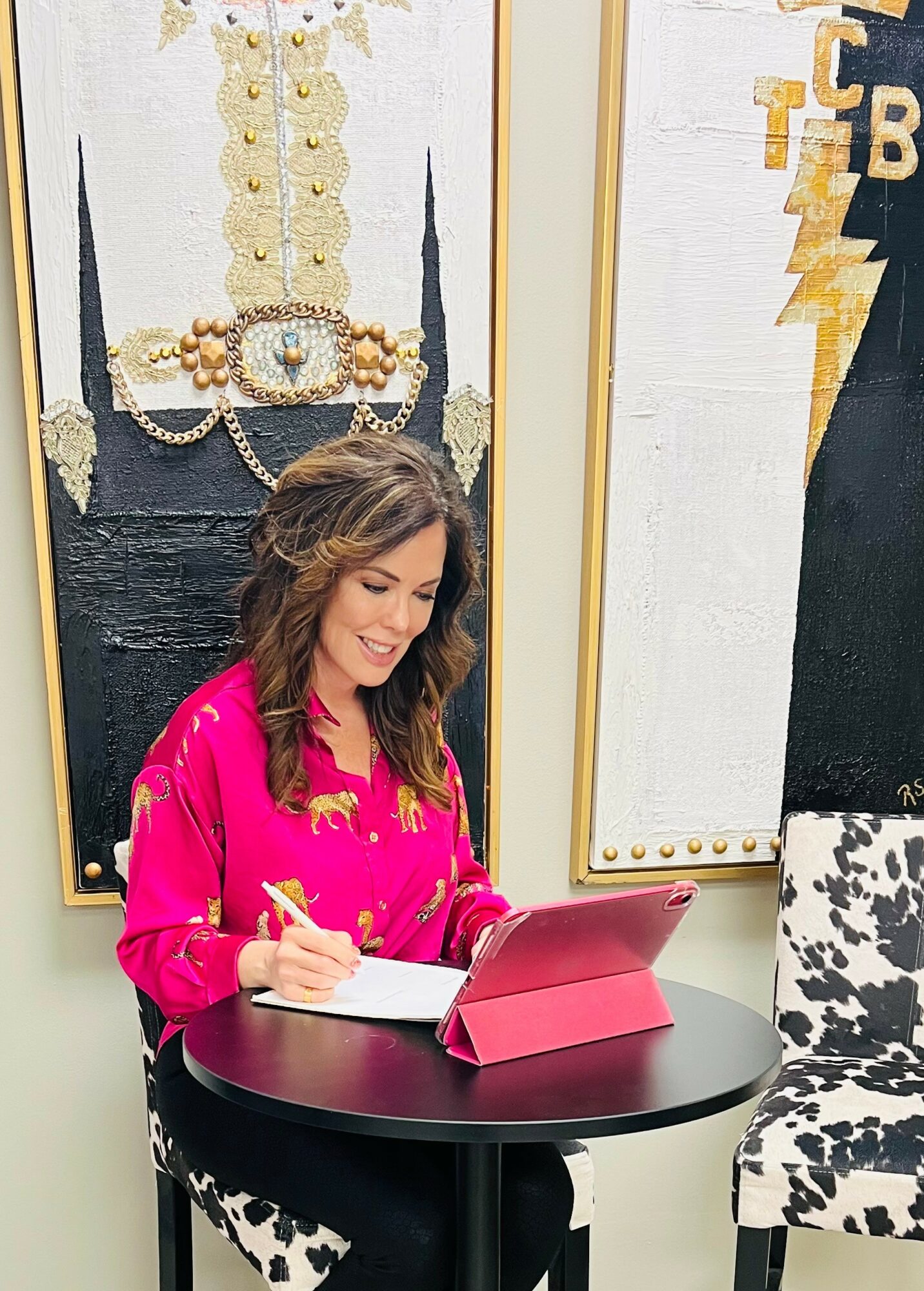 Woman with wavy hair in a pink blouse sitting at a round table with a pink tablet, smiling and writing.
