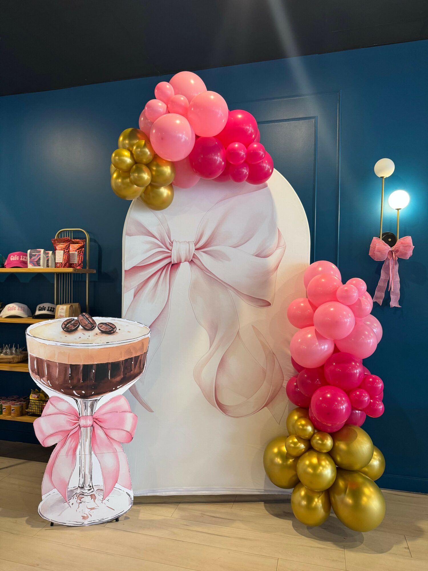 Decorative display with pink, gold, and pink balloons, a cake with a pink ribbon, and a backdrop with a bow design.