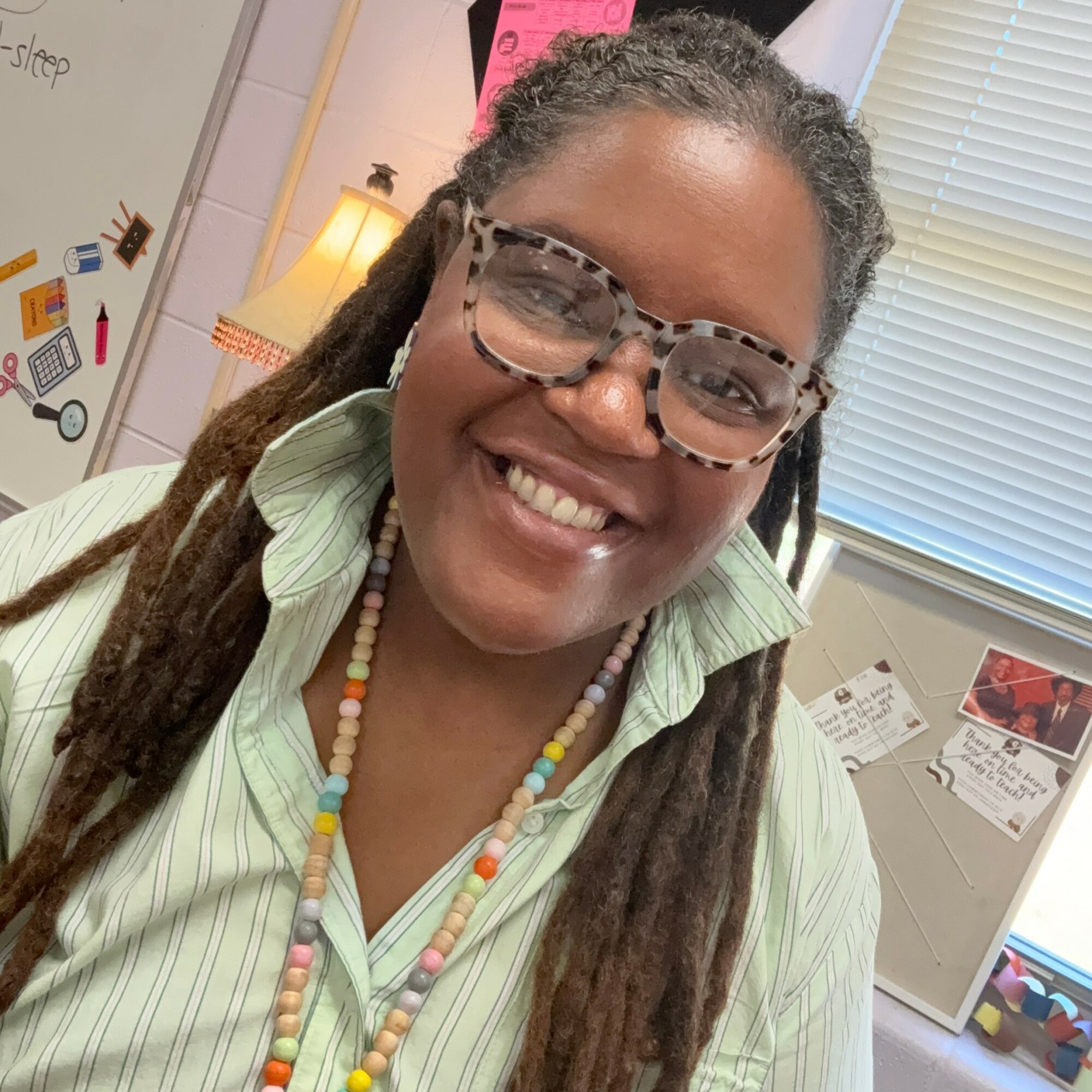 Smiling woman with glasses and dreadlocks, wearing a light green striped shirt, in an office setting.