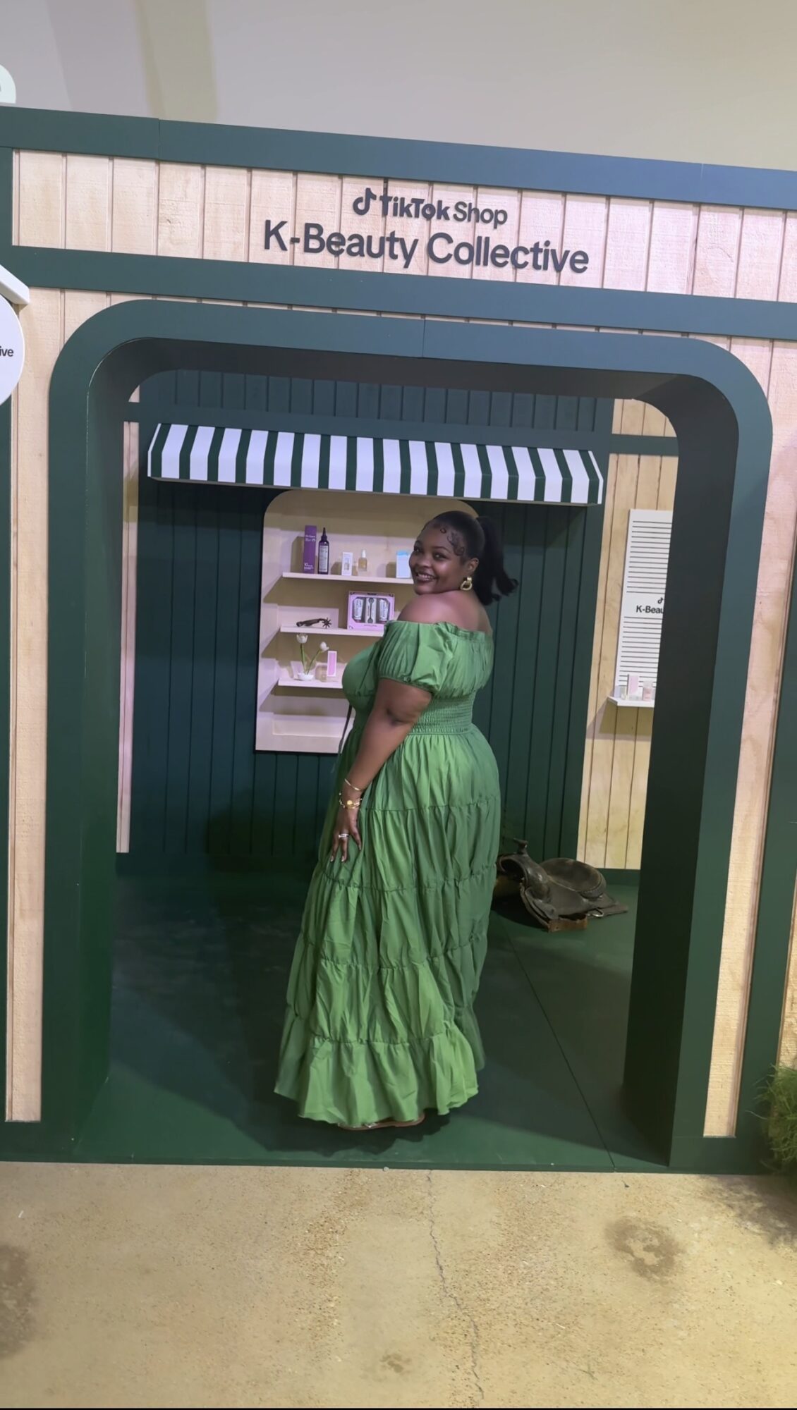 Woman in green dress standing inside a small shop with shelves and a sign above.