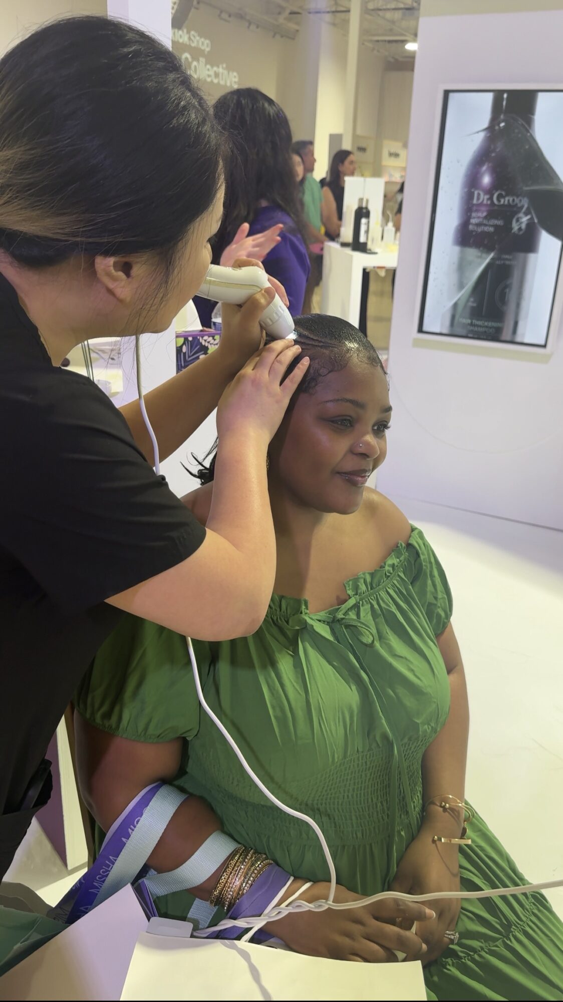 Woman receiving a cosmetic procedure on her forehead at a beauty event, with a display screen in the background.