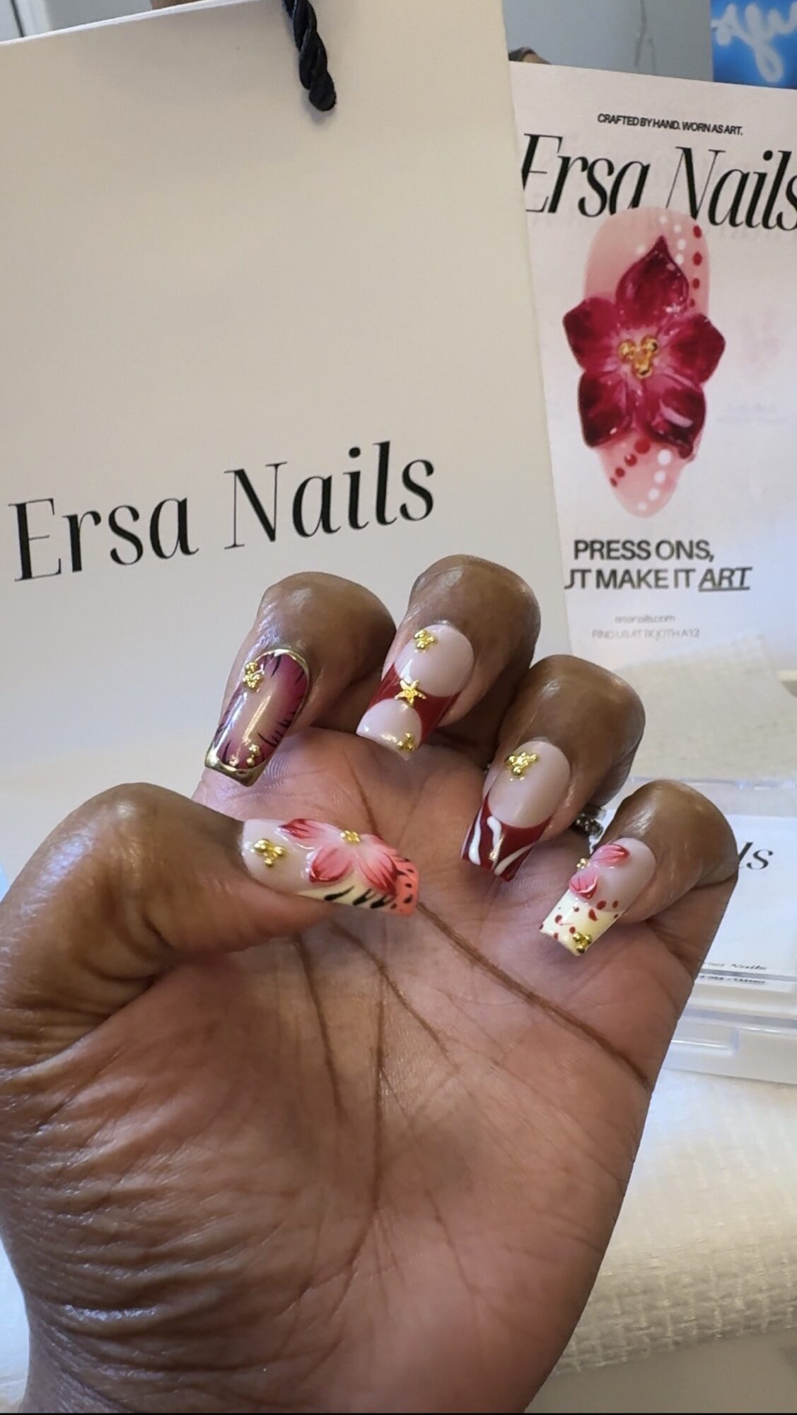 Hand with decorated nails featuring pink, white, and gold designs, holding a white shopping bag with black text.