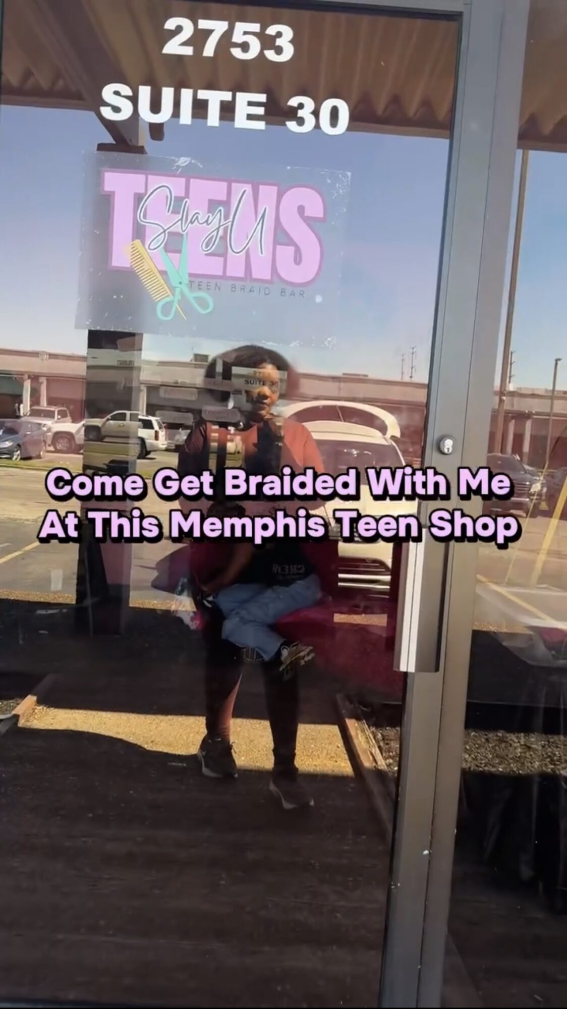 Reflection of a person taking a photo in a glass door with text and a store sign, with parking lot outside.
