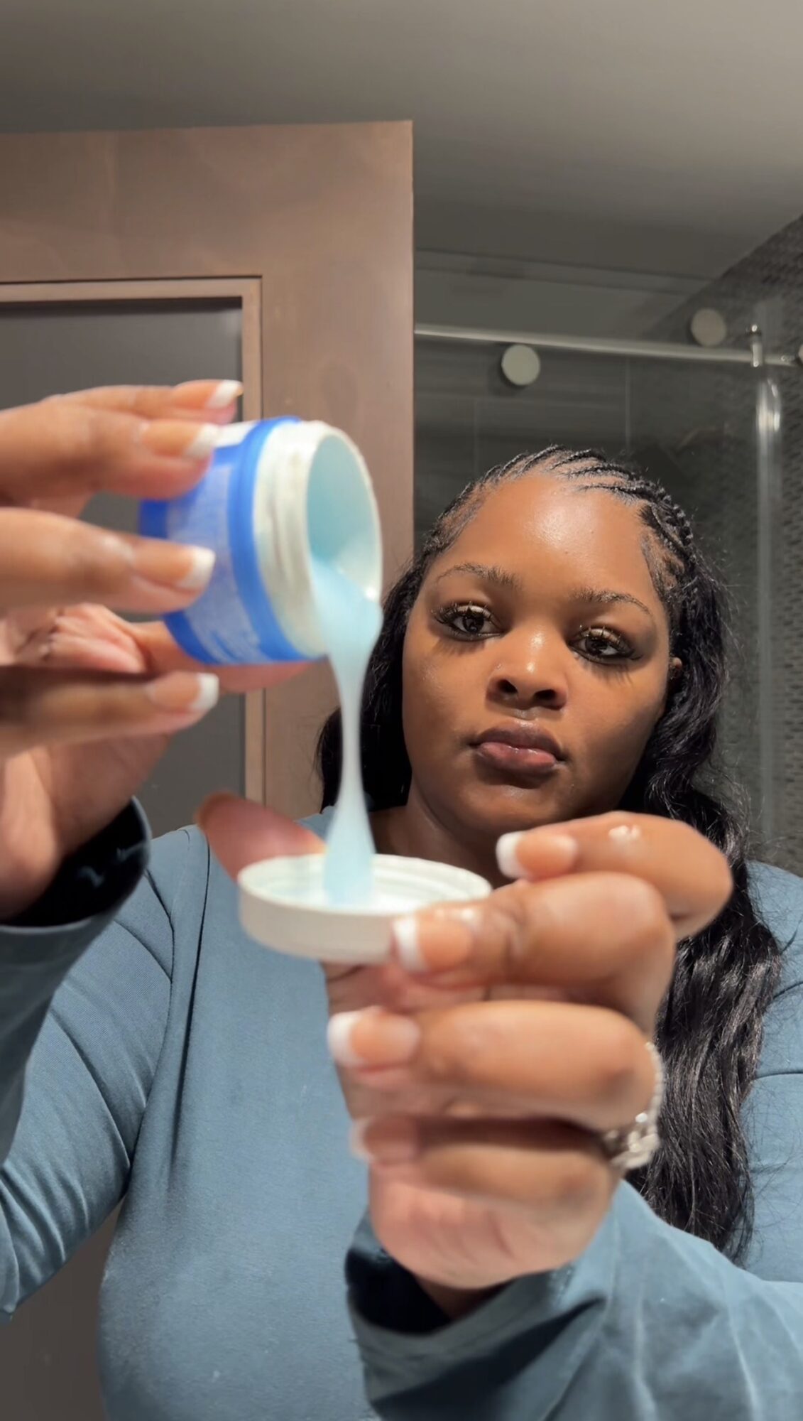 Woman pouring blue liquid from container into small dish, holding it with both hands, in a room with gray walls.