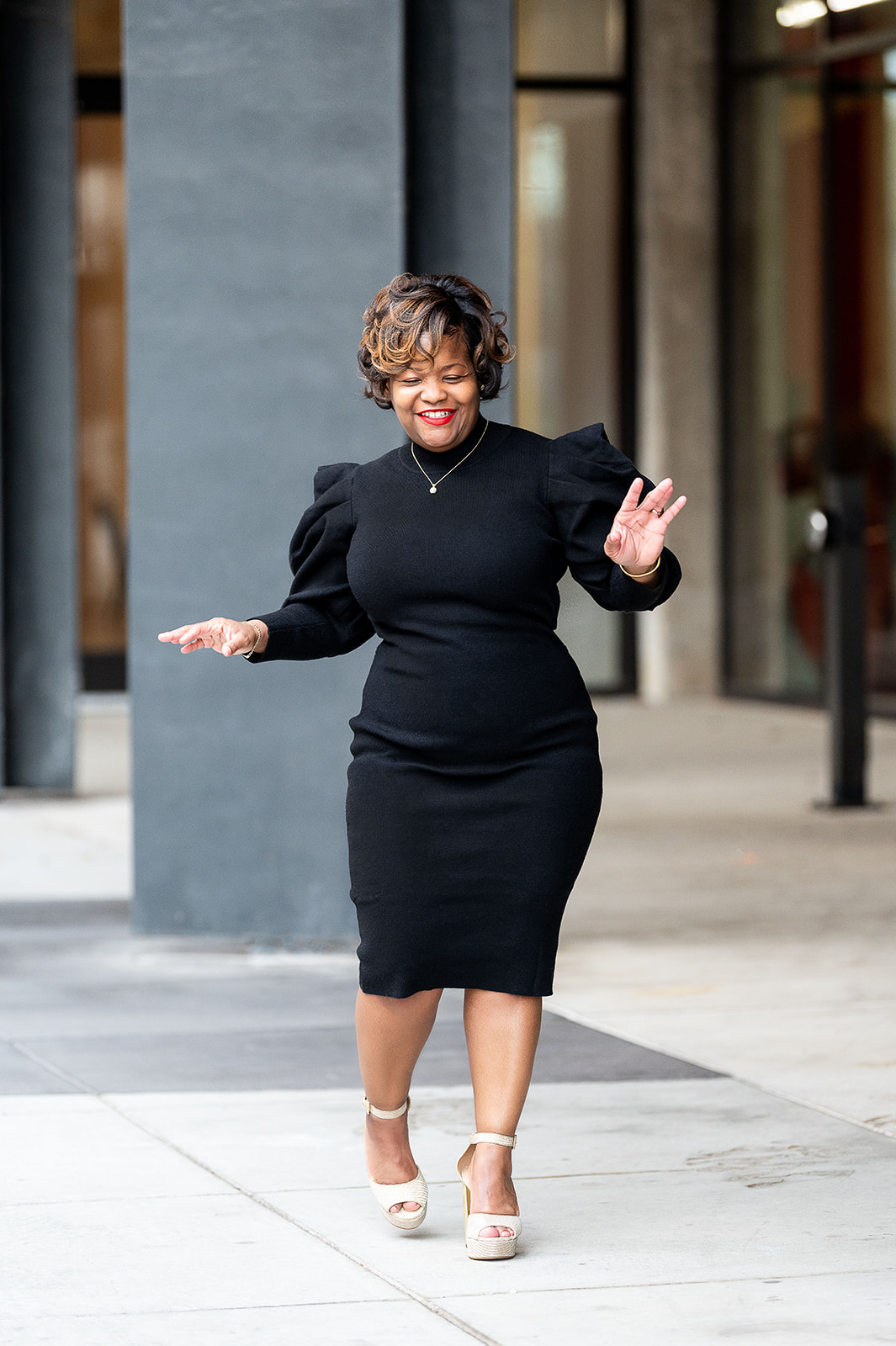 Woman in black dress walking outdoors, smiling, with buildings in background.