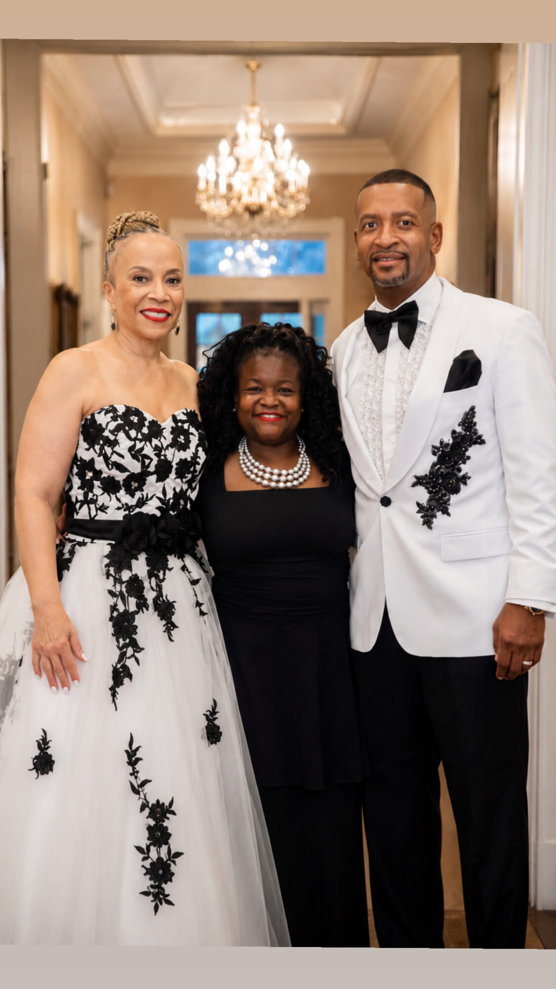 Three people dressed formally, standing in a hallway with a chandelier overhead, smiling at the camera.