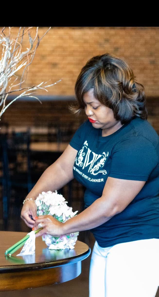 Woman with short hair arranging flowers on a table, brick wall background, indoors.