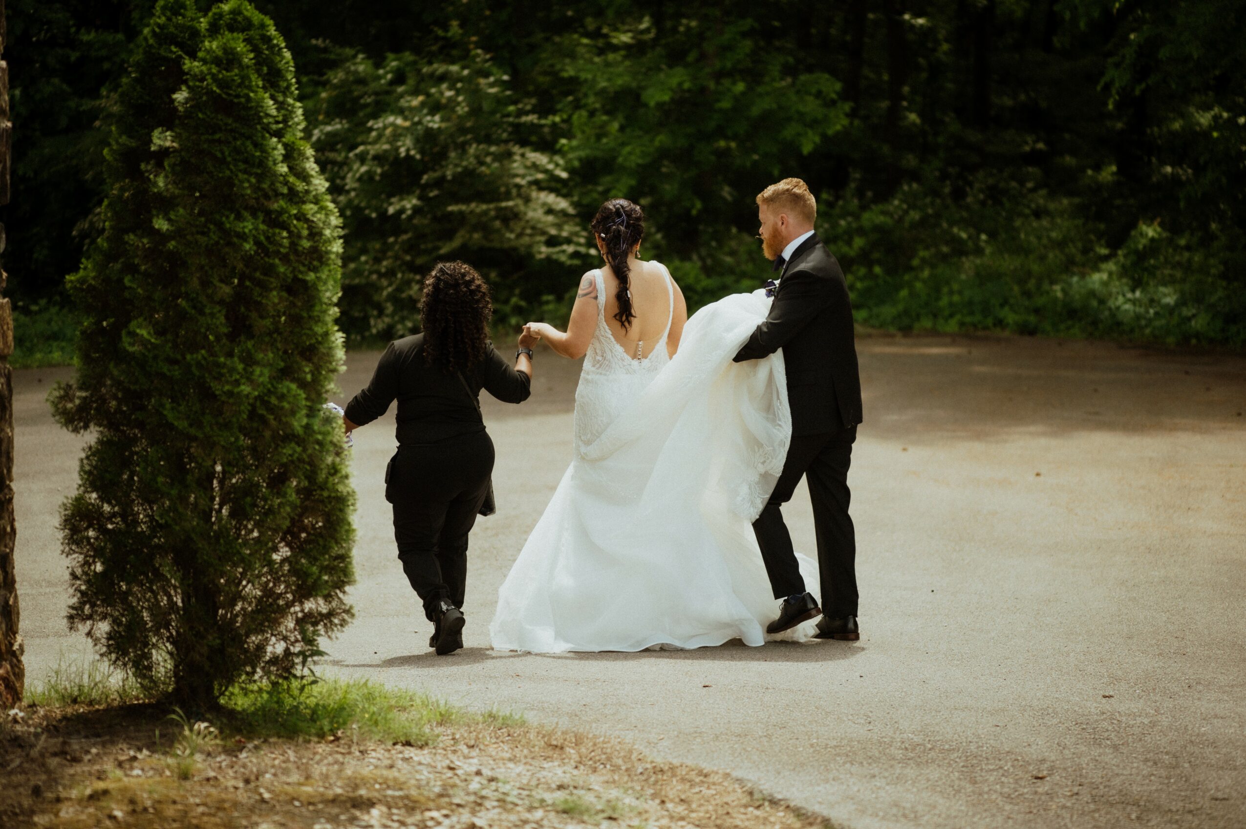 Three people, two women and one man, walk together outdoors on a dirt path surrounded by trees and greenery.