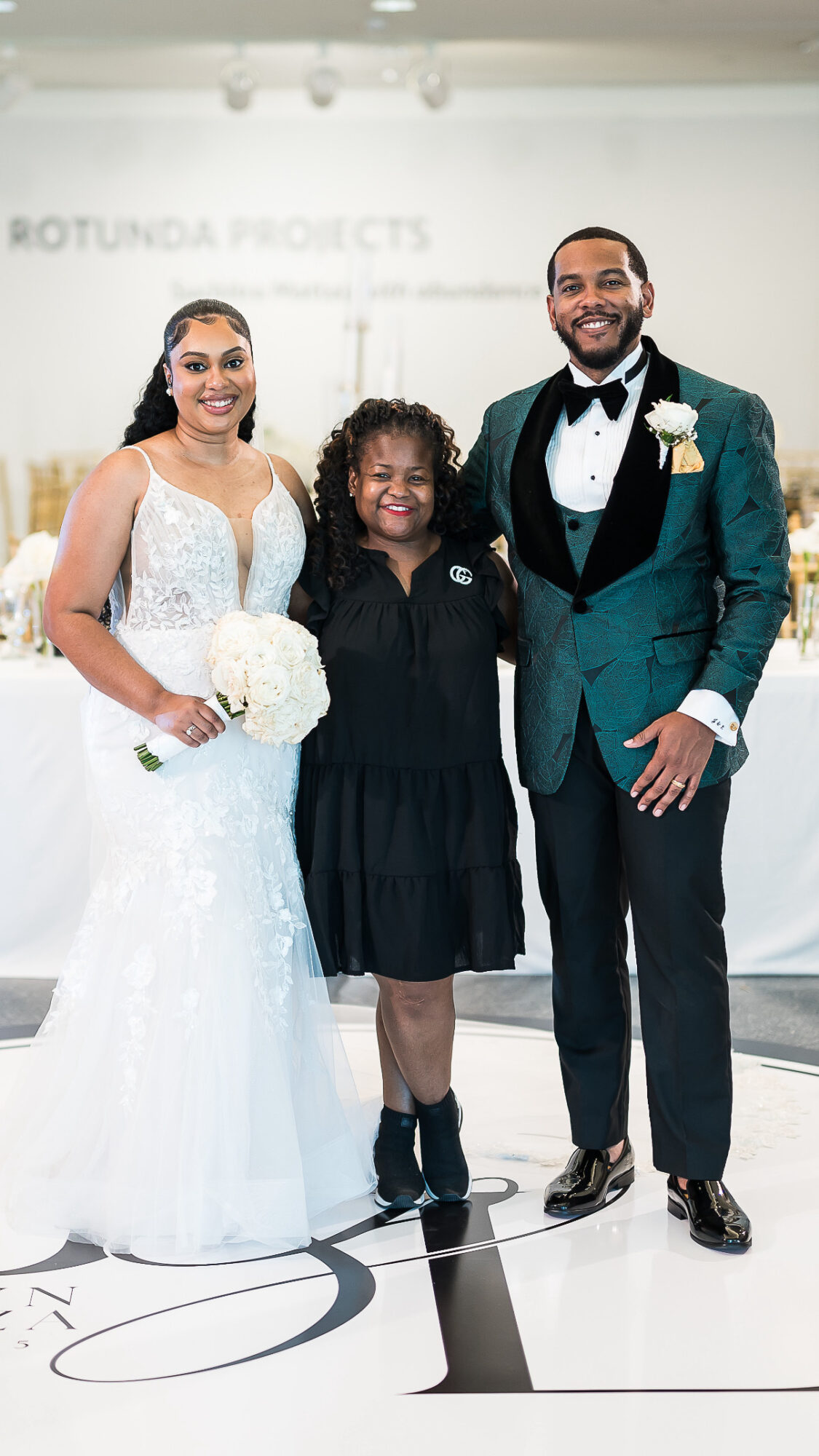Three people standing together at a wedding reception, smiling, with a white background and a blurred sign behind them.
