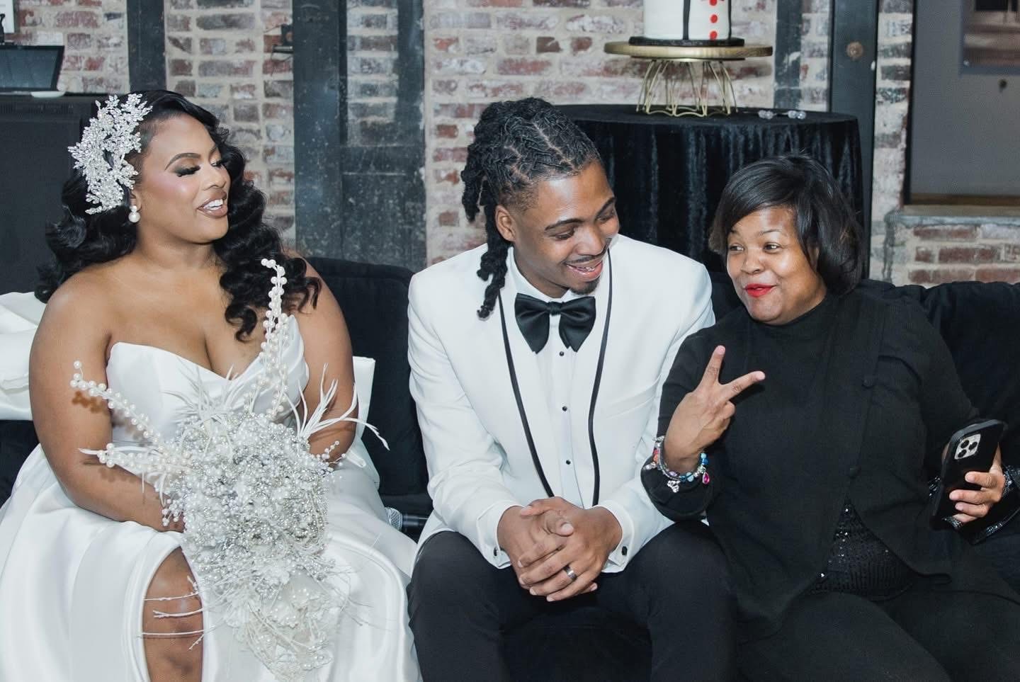 Three women sitting on a couch, one in a wedding dress, two in formal attire, smiling and talking, brick wall background.