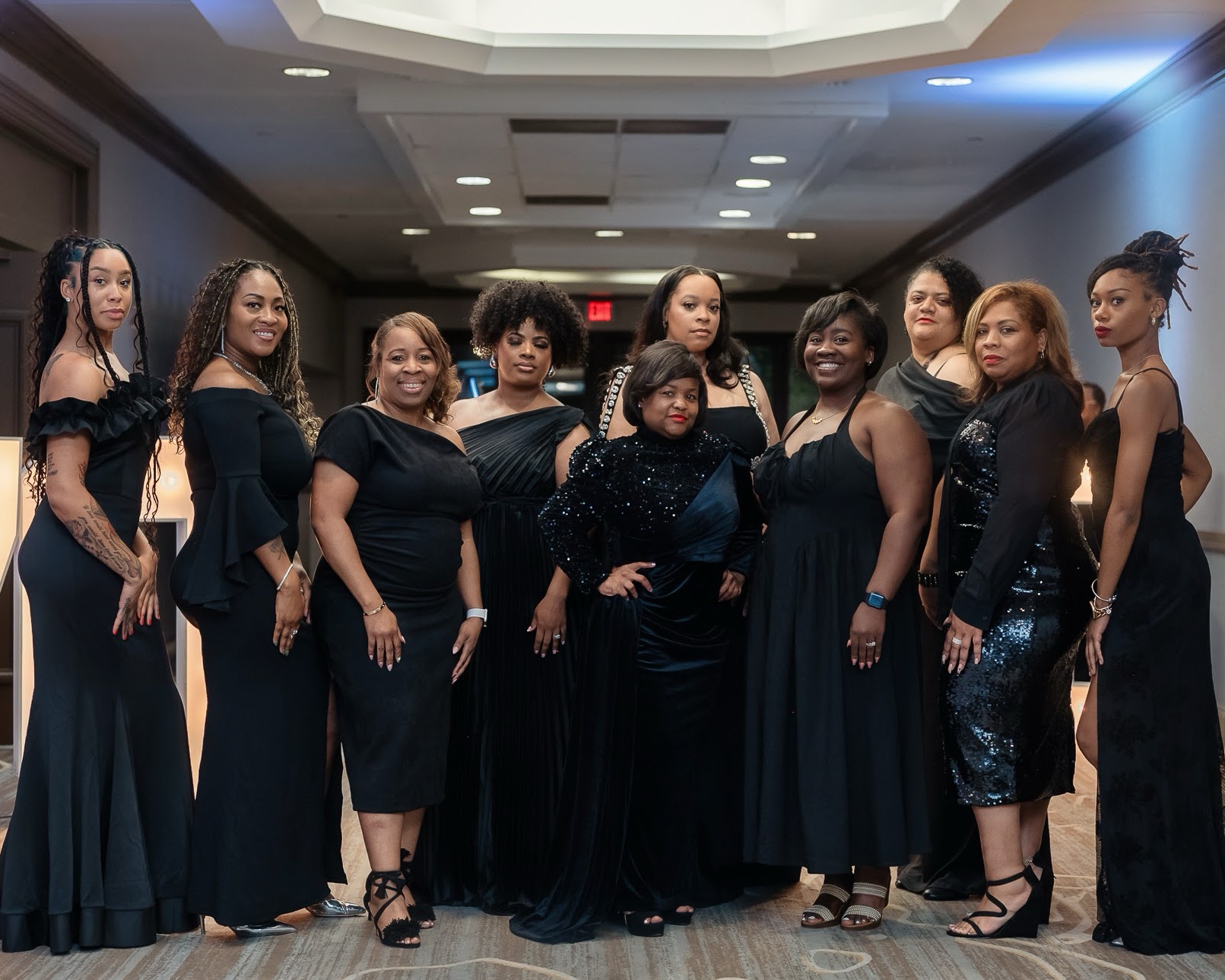 Group of women in black dresses standing together in a room with ceiling lights and a hallway background.