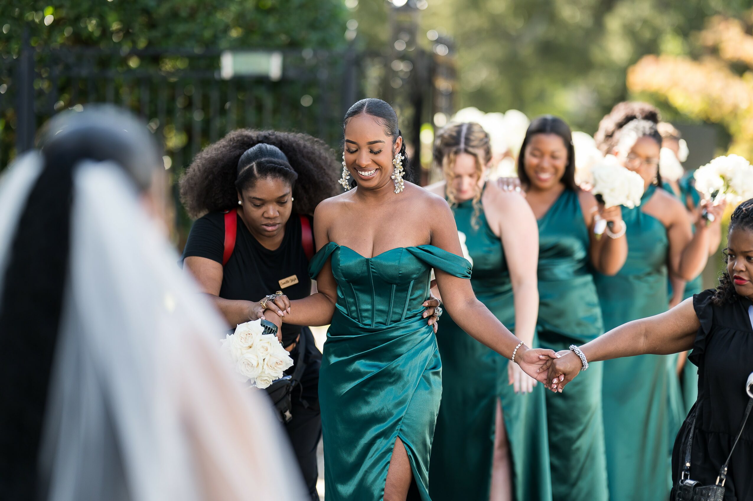 Group of women in teal dresses holding hands outdoors, smiling, with trees in the background.