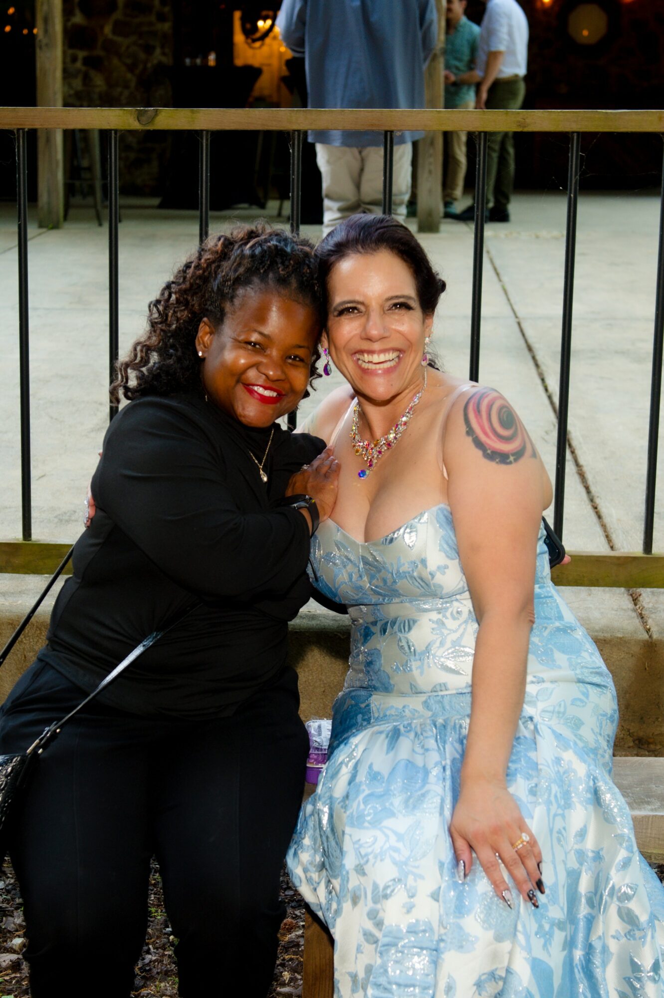 Two women smiling, one in a black outfit and the other in a blue dress, sitting close together outdoors.