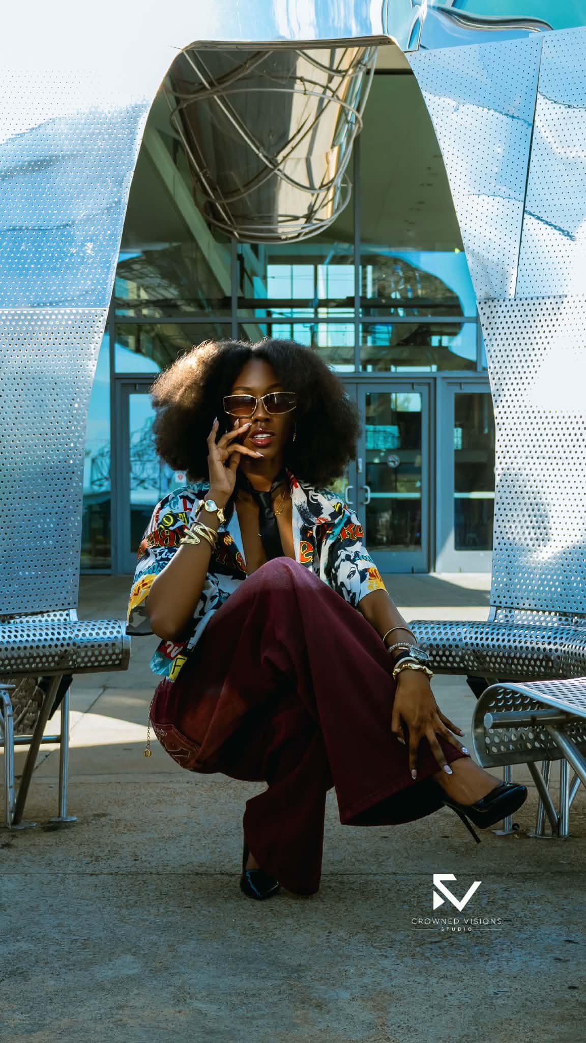Woman with sunglasses and curly hair sitting in front of a mirror, wearing colorful clothing and jewelry.
