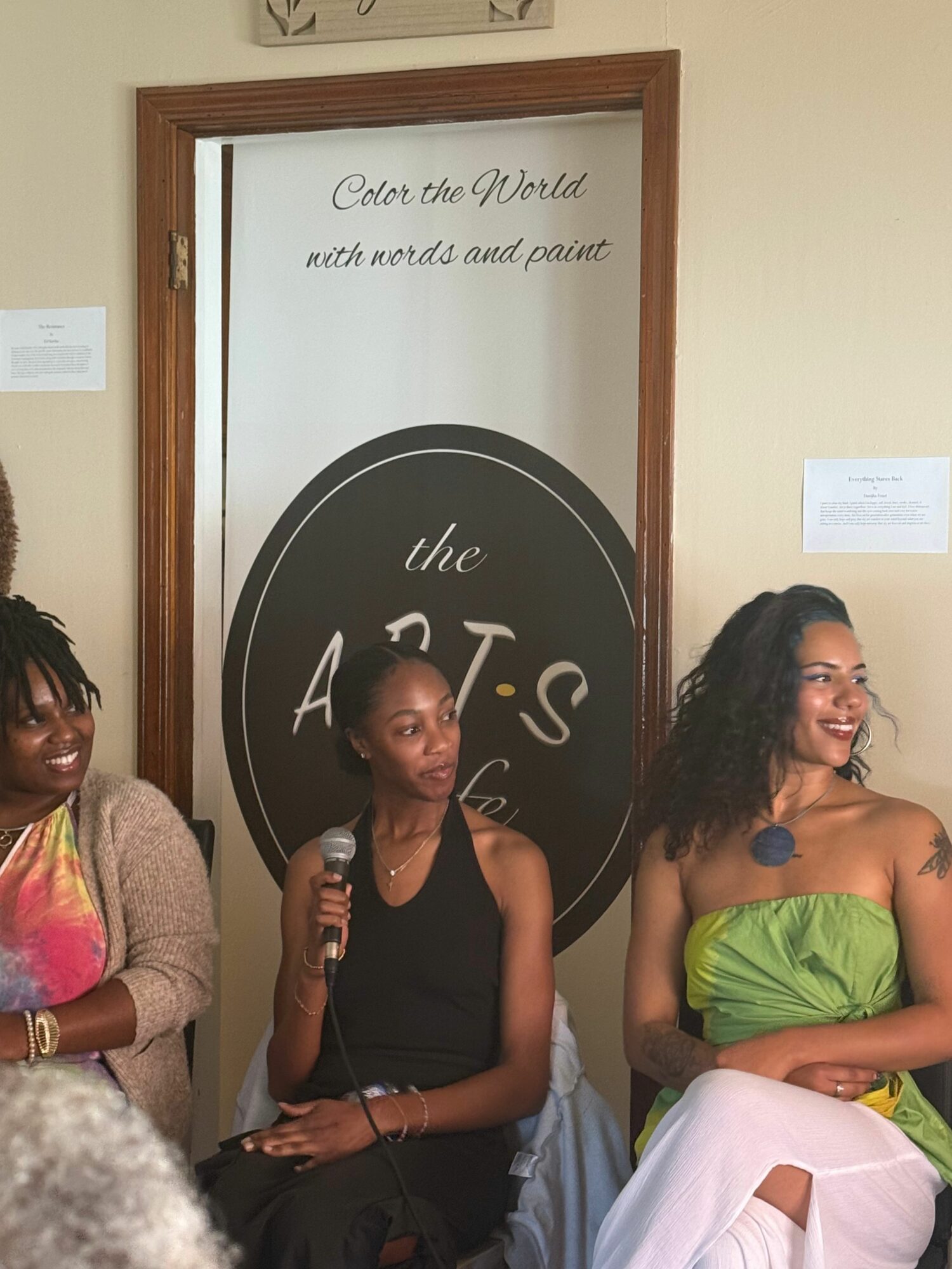 Two women sitting in front of a sign that reads 'the ART'S'. One woman is wearing a green top and white pants, the other is in a colorful dress, holding a microphone.