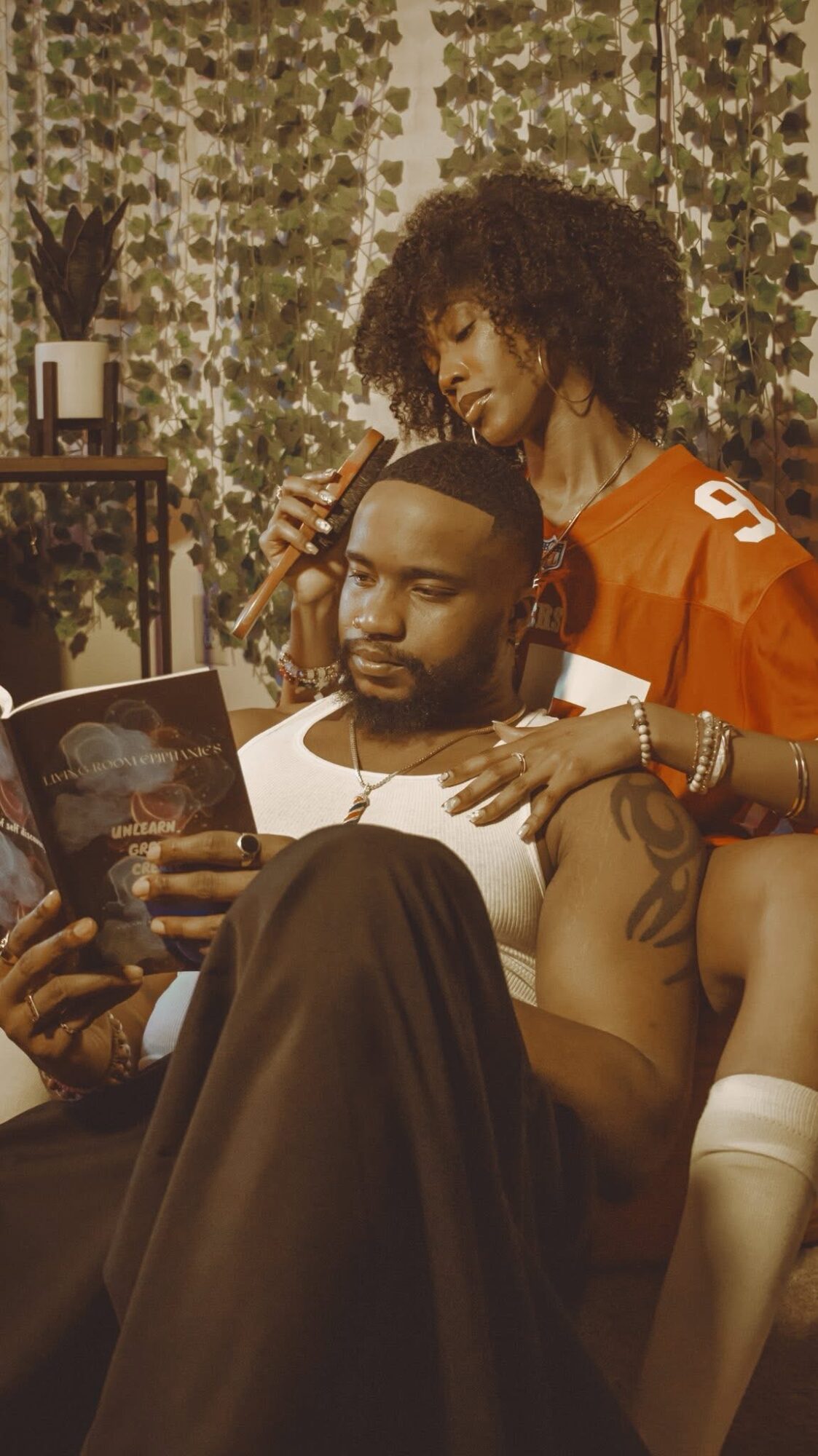 Two women and a man in a cozy room with plants and warm lighting, one woman reading a magazine, the other styling hair.