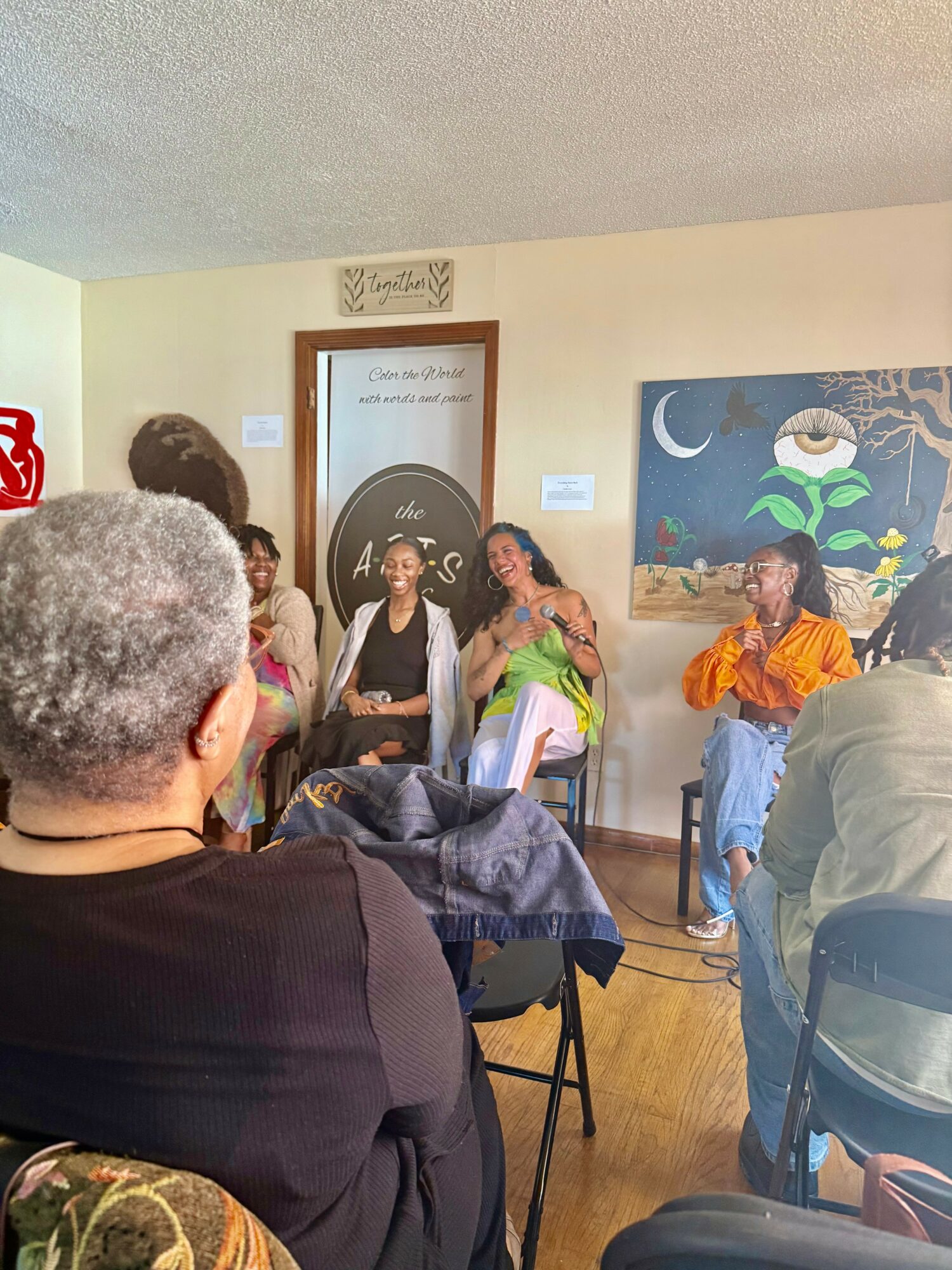 Group of people sitting and talking in a room with artwork on the wall, some with microphones, engaging in discussion.