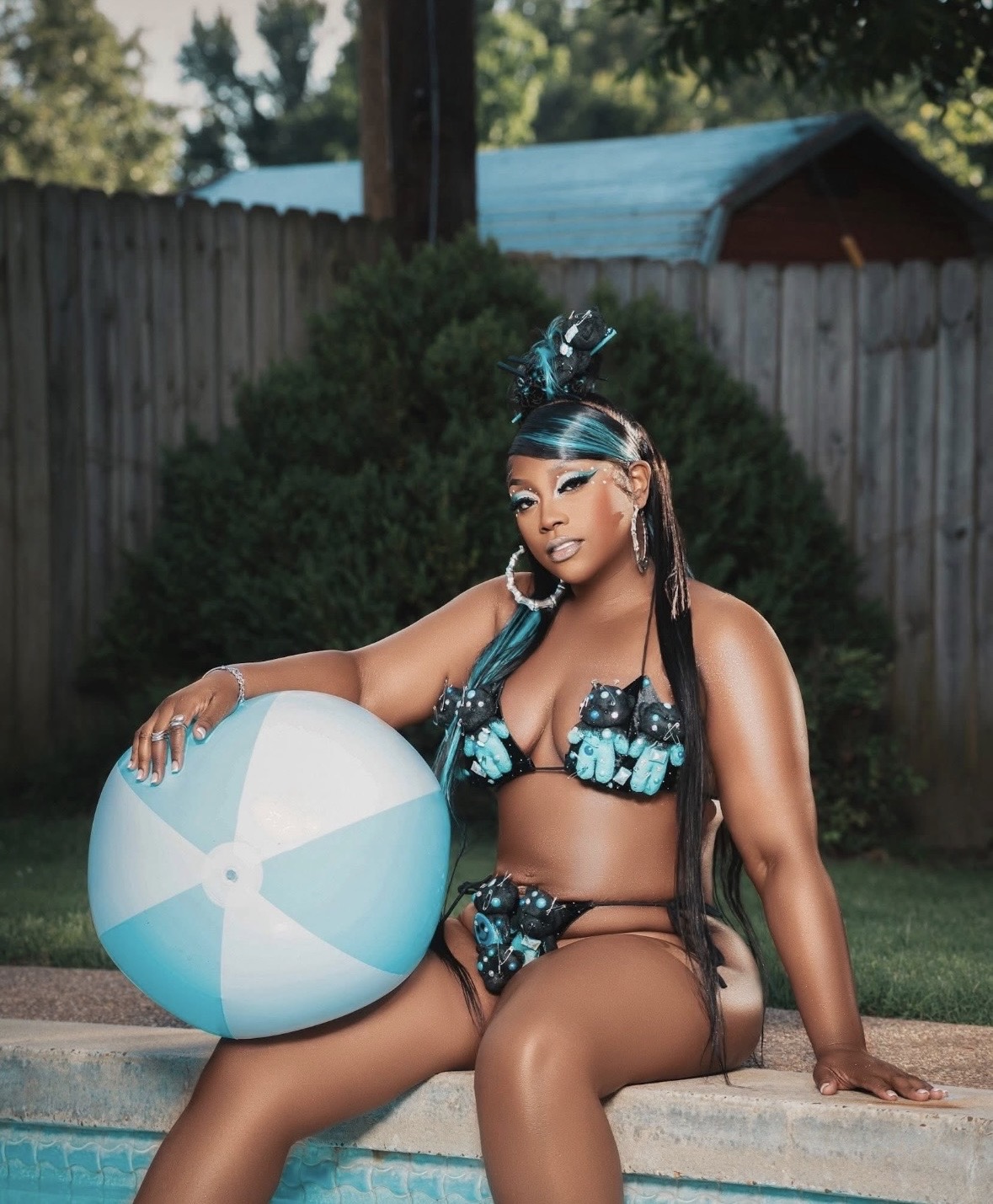 Woman in a decorated bikini sitting by a pool with a beach ball, outdoor setting, fence and trees in background.