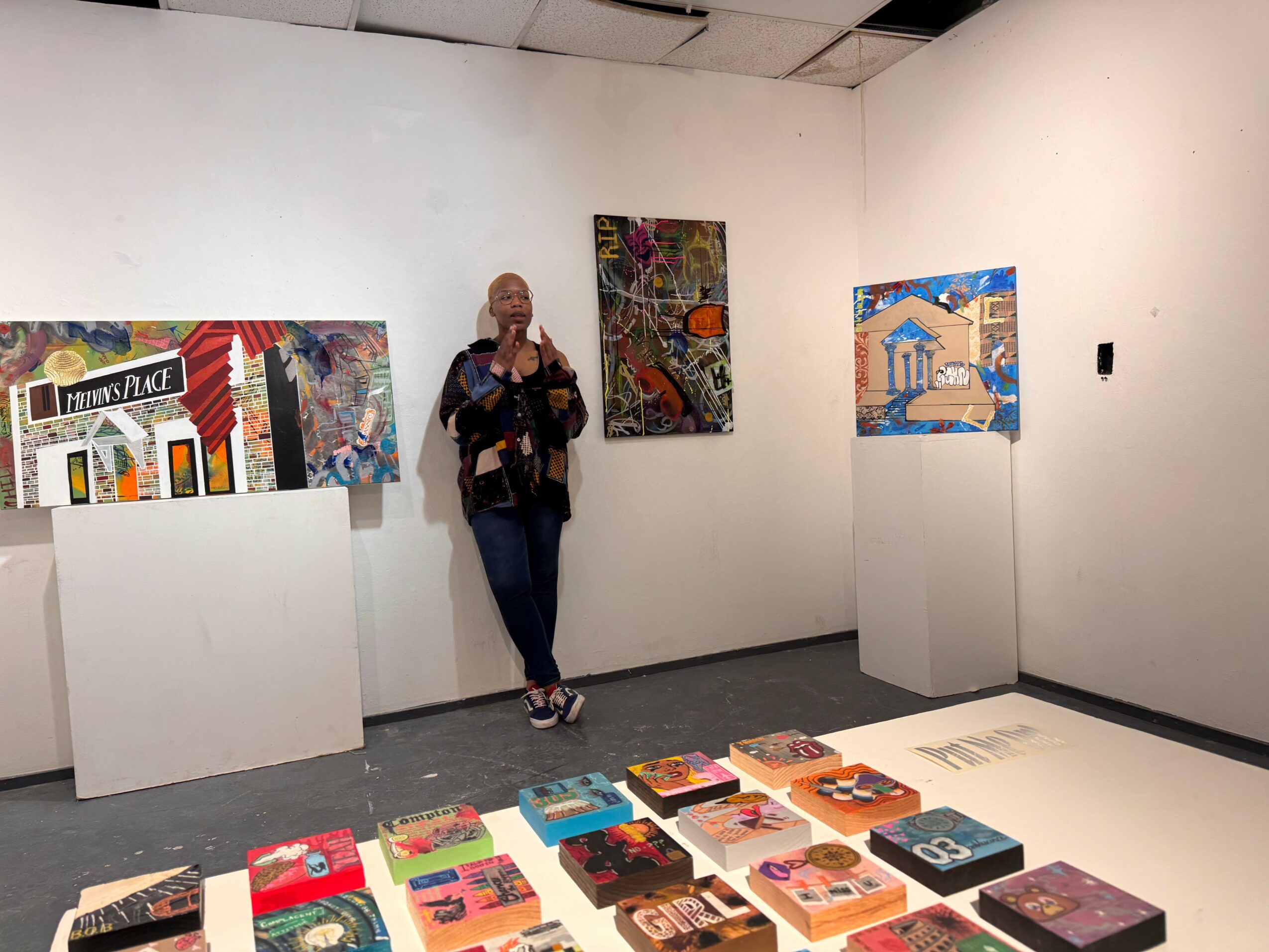 Person taking a photo in an art gallery with paintings on white walls and artwork on display, floor with books or catalogs.