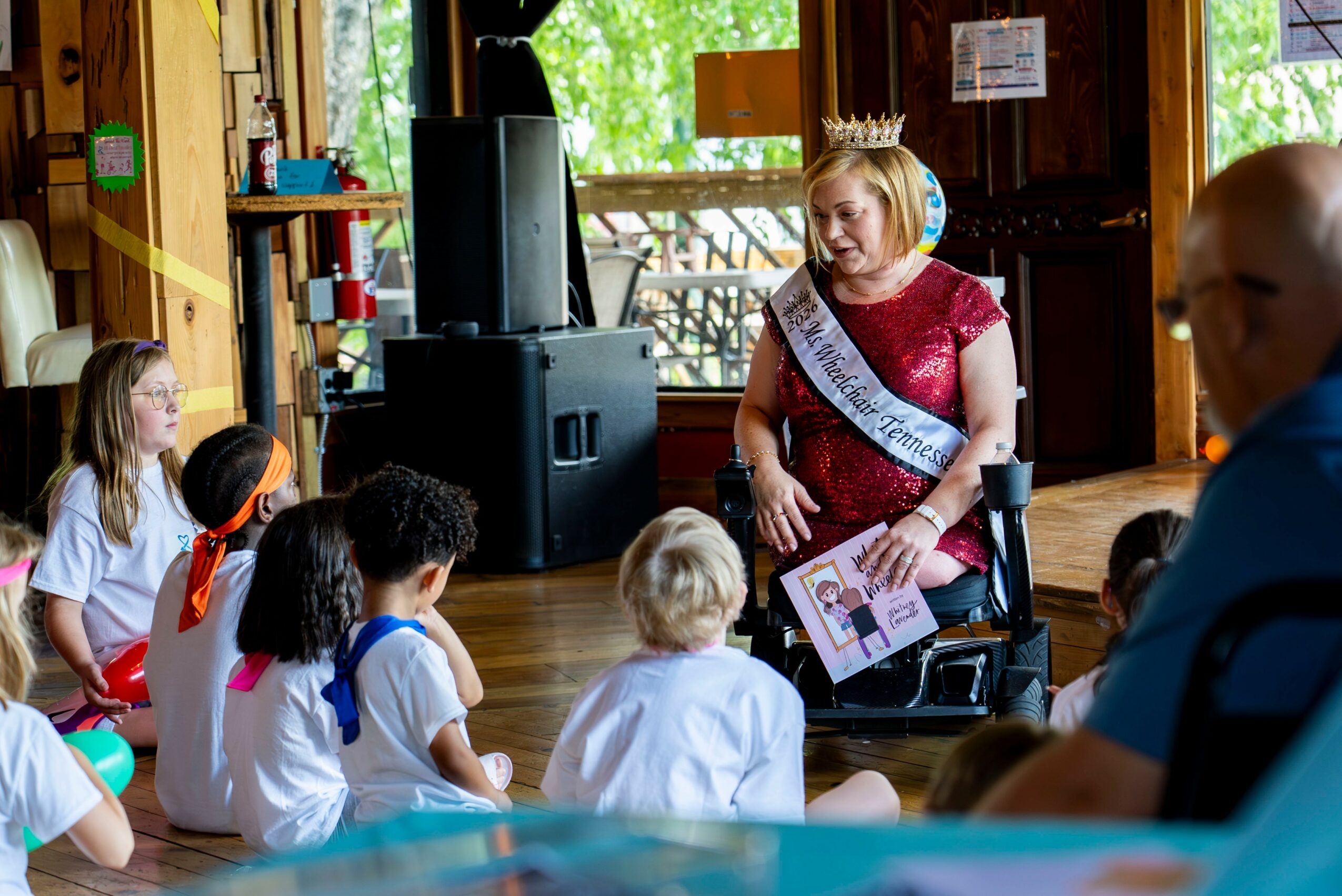 Woman wearing a sash and crown speaks to children sitting on the floor in a wooden room with large windows.