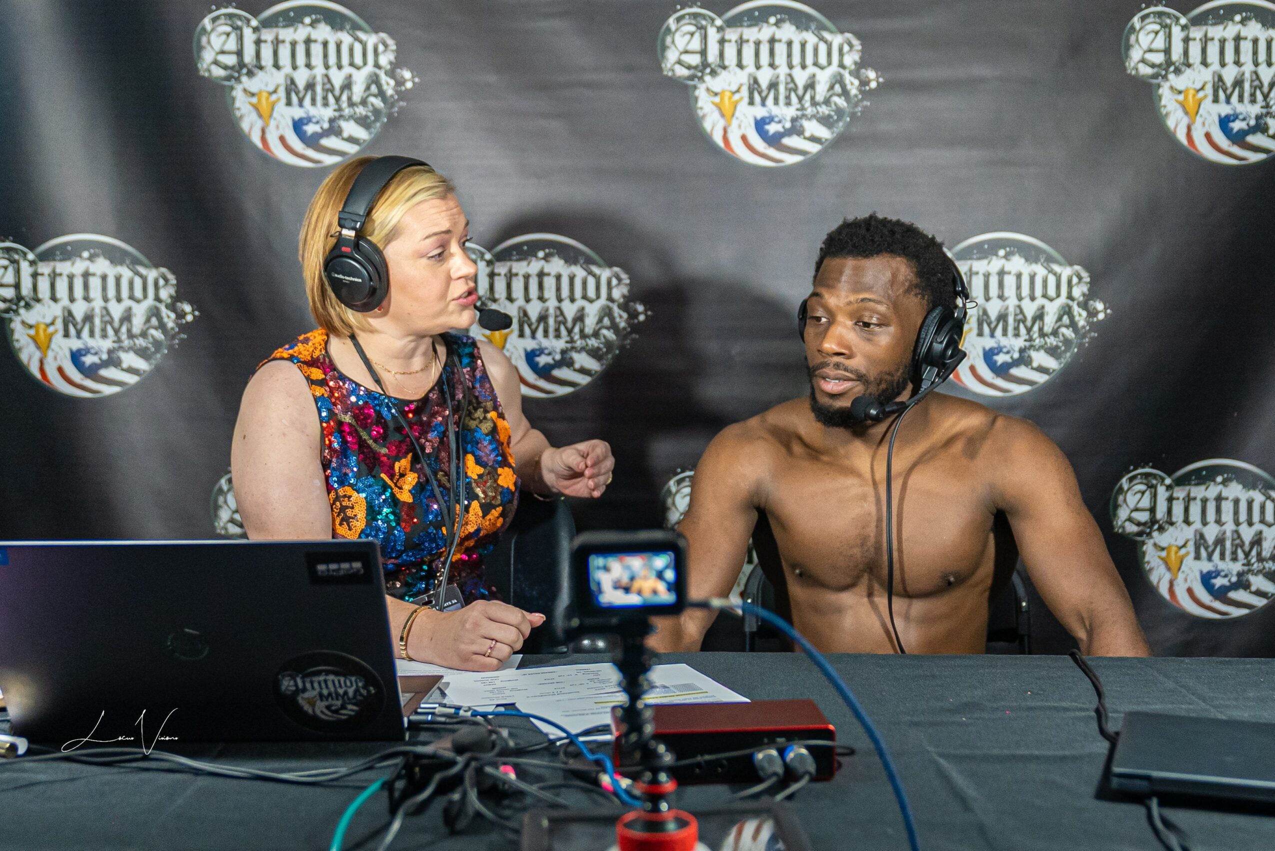 A woman and a shirtless man with headphones sit at a table with recording equipment, in front of a backdrop with logos.