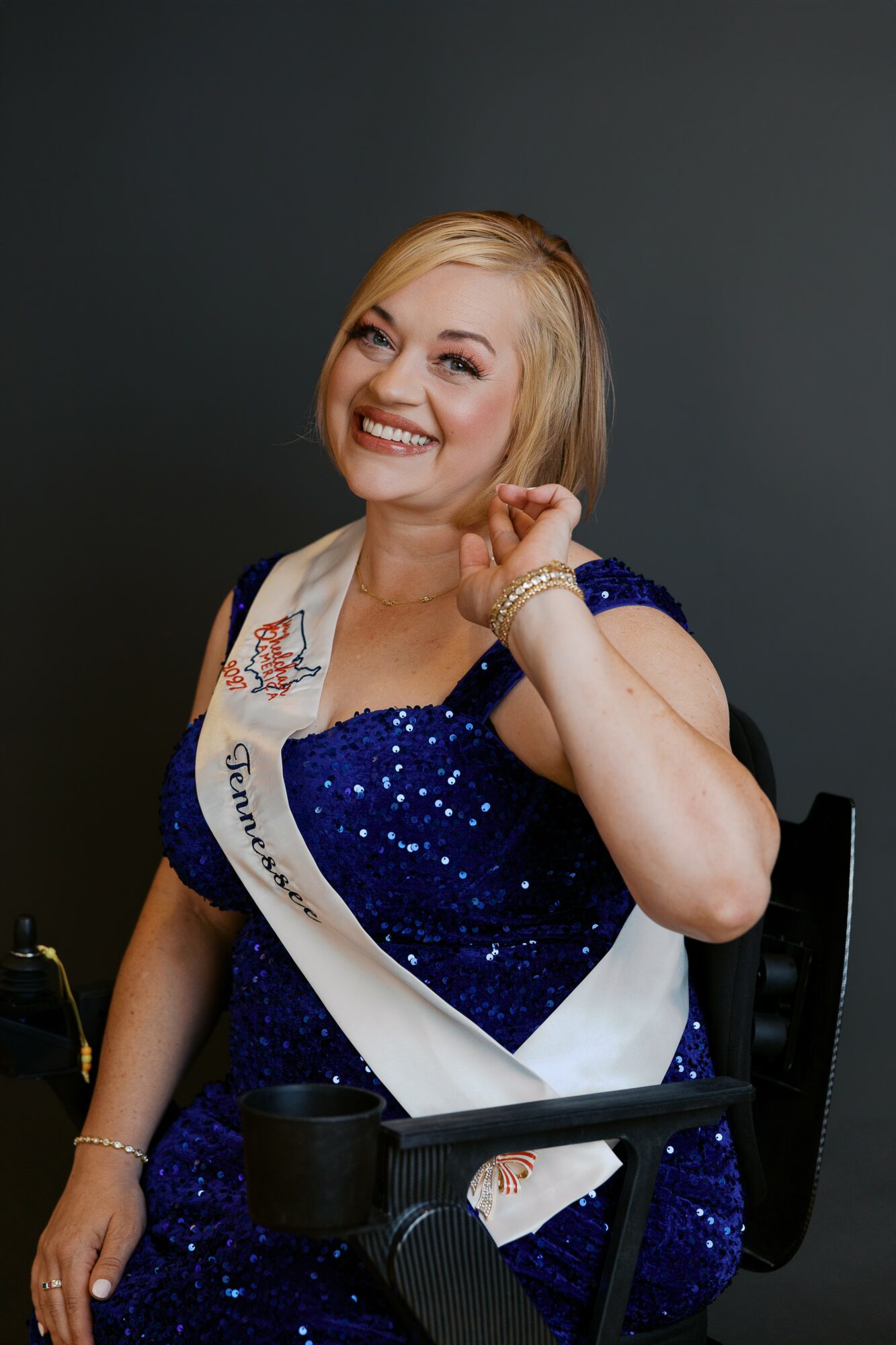 Woman smiling, wearing a blue sequined dress and sash, sitting in a chair against a dark background.