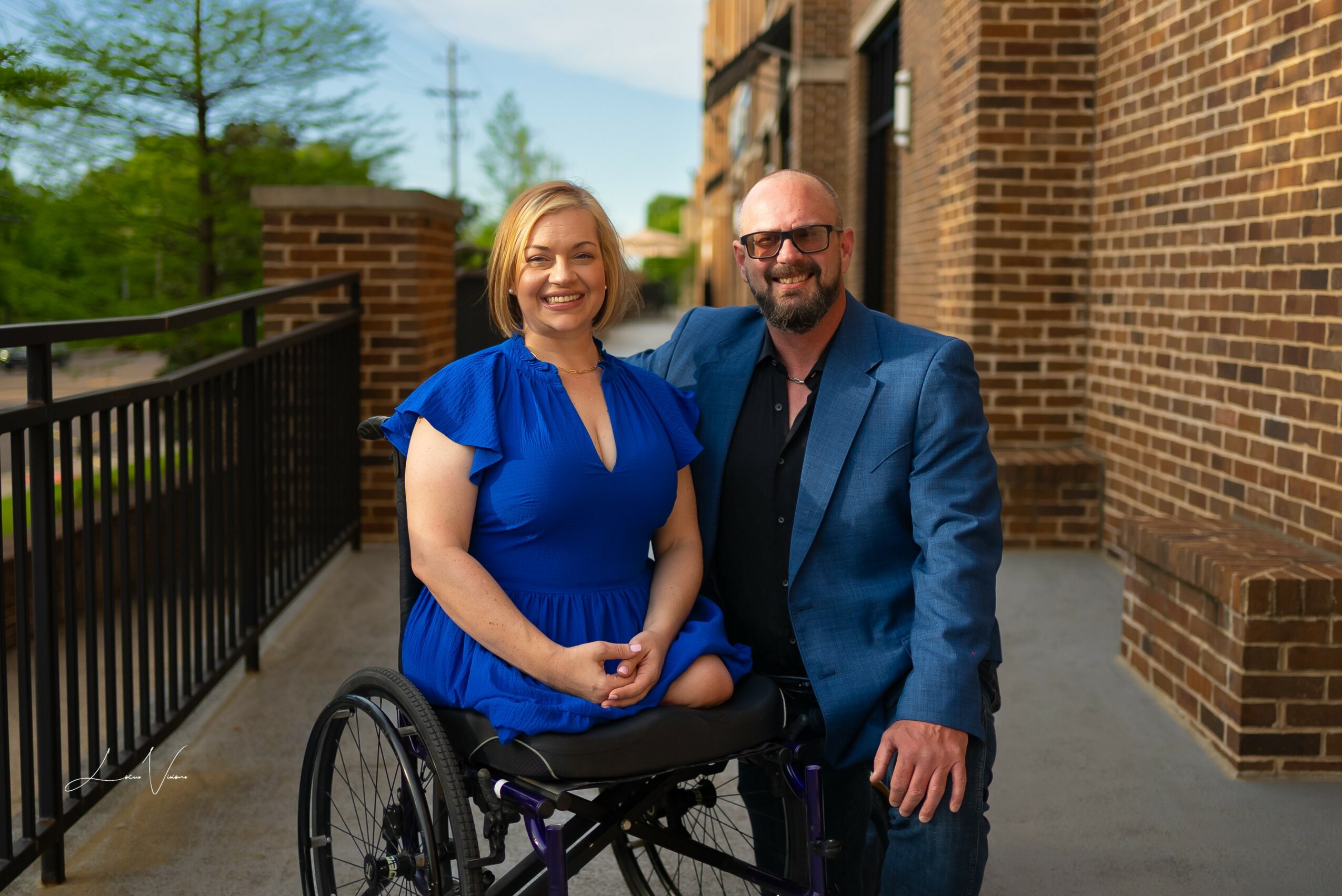 A woman in a blue dress and a man in a blue suit sit together outdoors, smiling, with a brick building in the background.