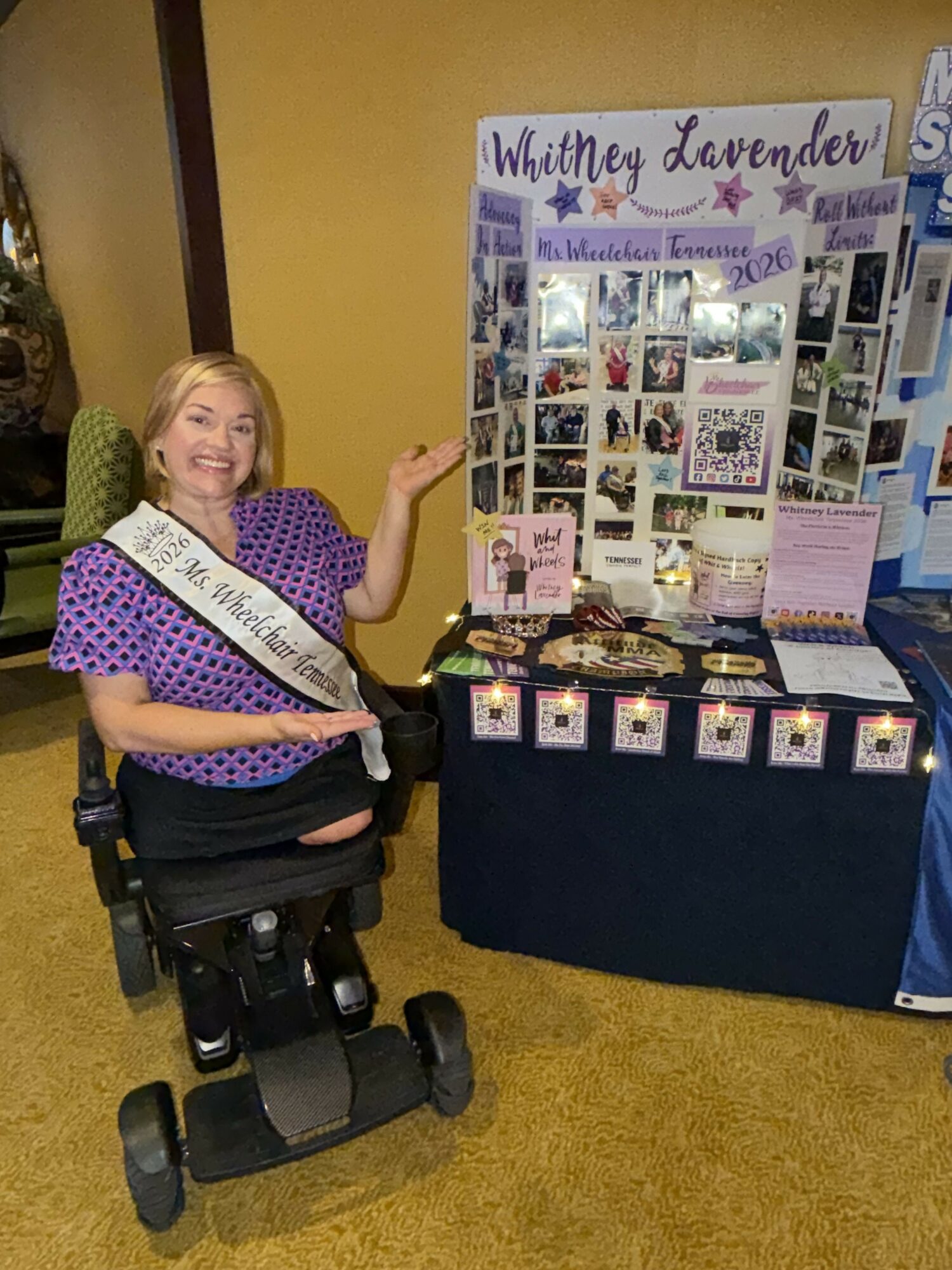 Woman sitting in a wheelchair in front of a display table with photos and informational materials, against a yellow wall.