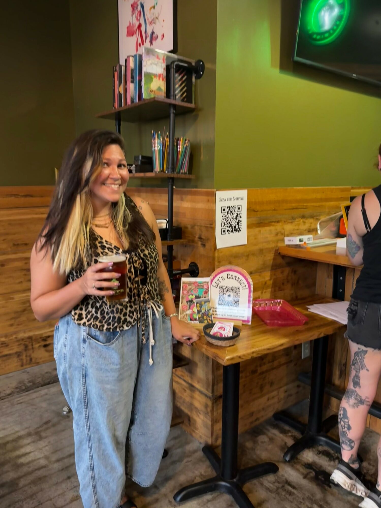 Woman with long dark hair holding a drink, standing near a wooden table in a room with green walls and bookshelves.