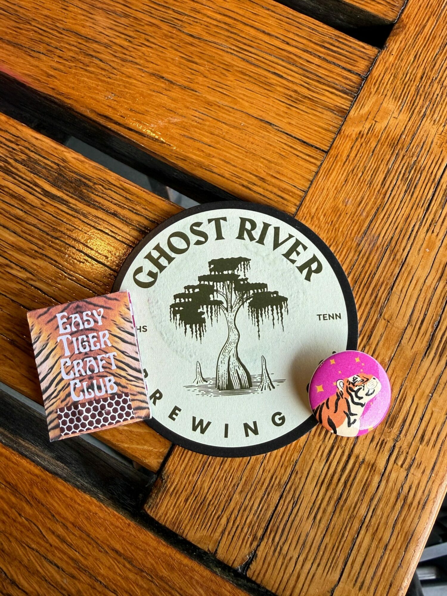 Round coaster with 'GHOST RIVER BREWING' text and a tree illustration, a pink and orange ball, and a small rectangular card on a wooden surface.