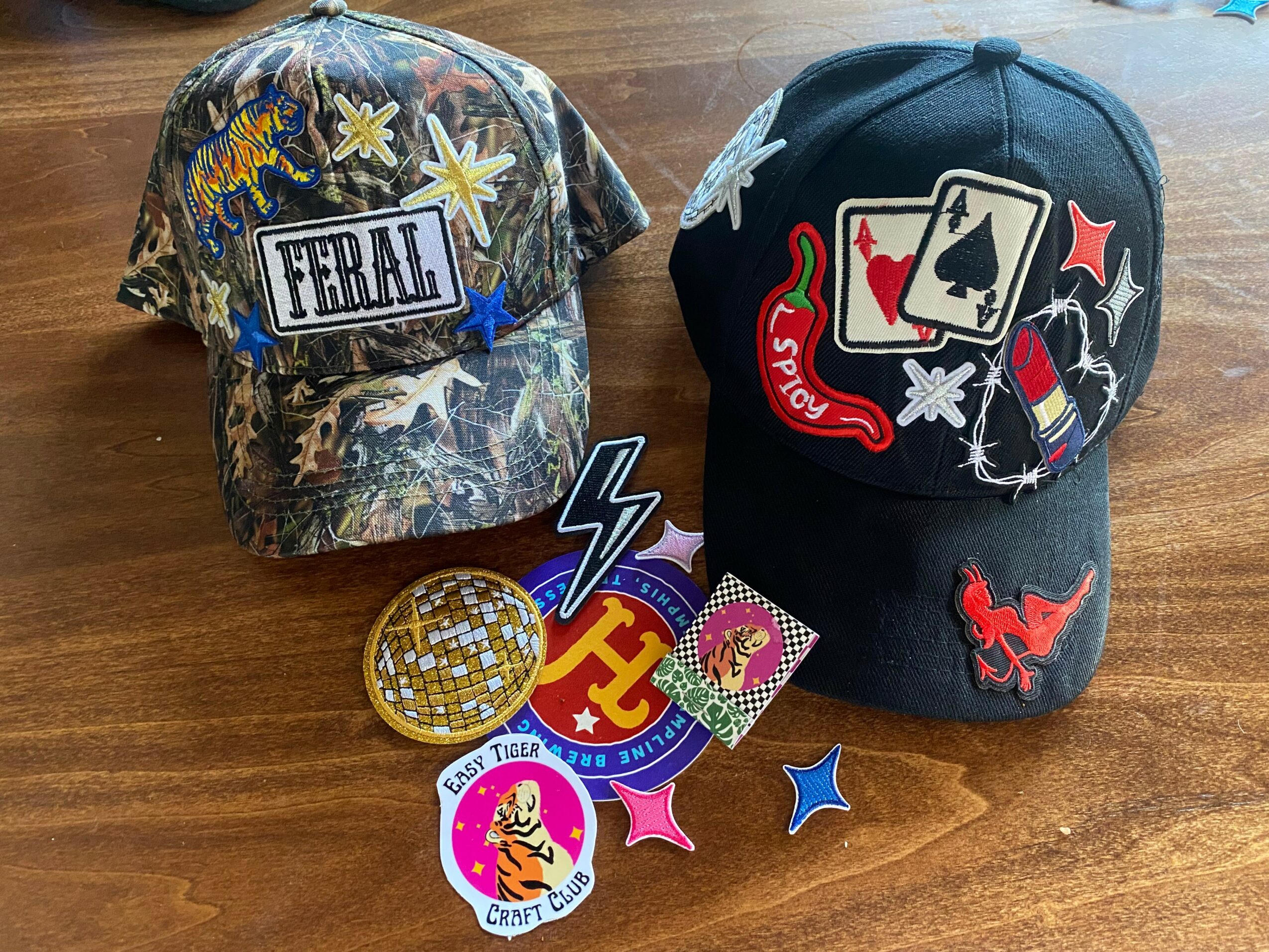 Two baseball caps, one with floral and patch design, the other with patches and embroidery, surrounded by pins and patches on a wooden surface.